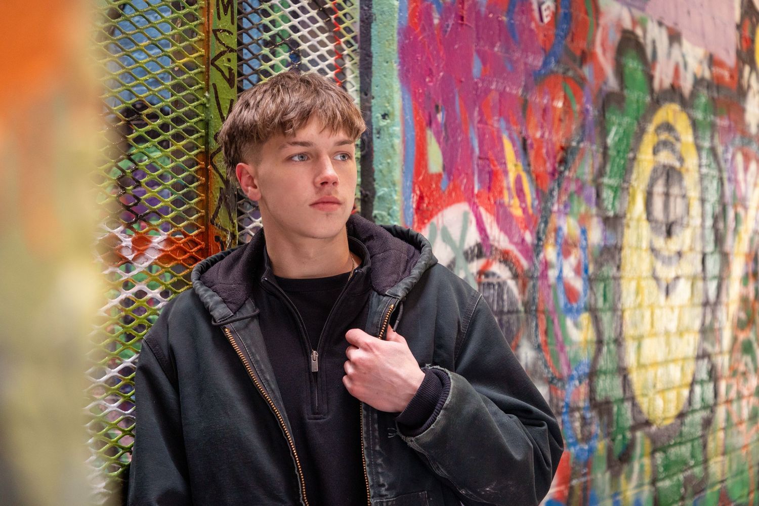 Person in dark jacket poses against colorful graffiti wall background.