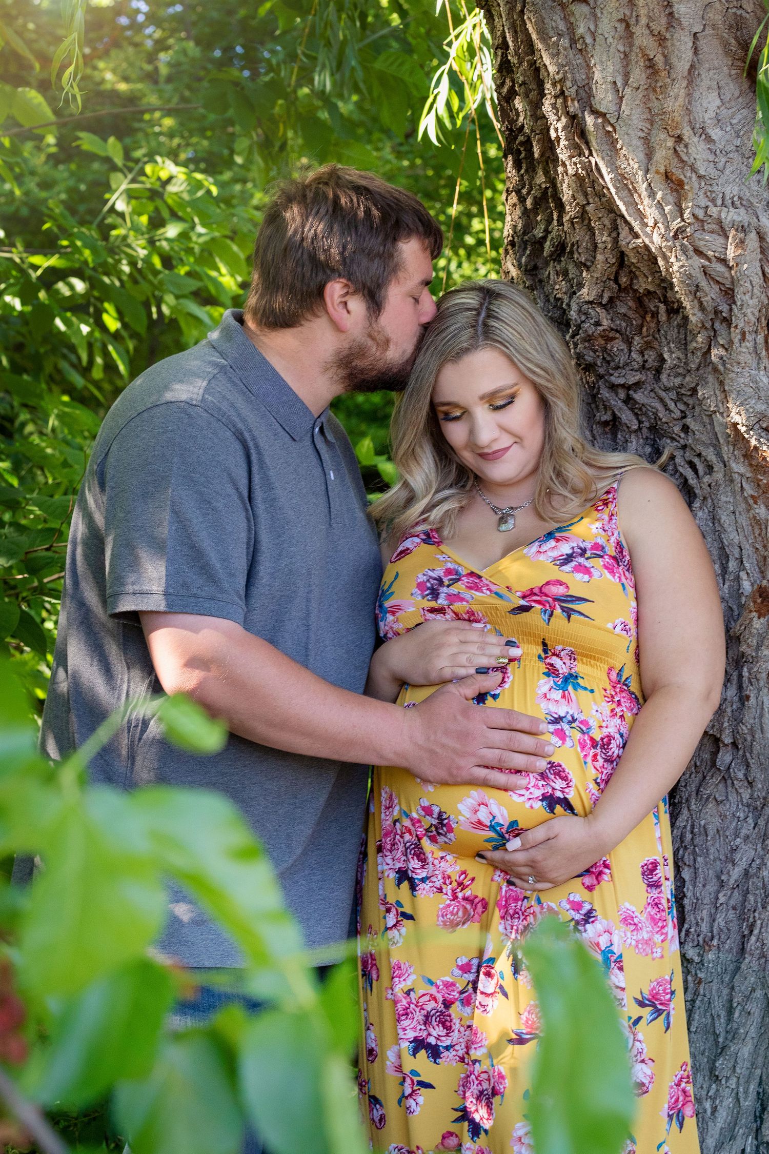 Romantic maternity photoshoot against a large tree trunk in dappled sunlight.