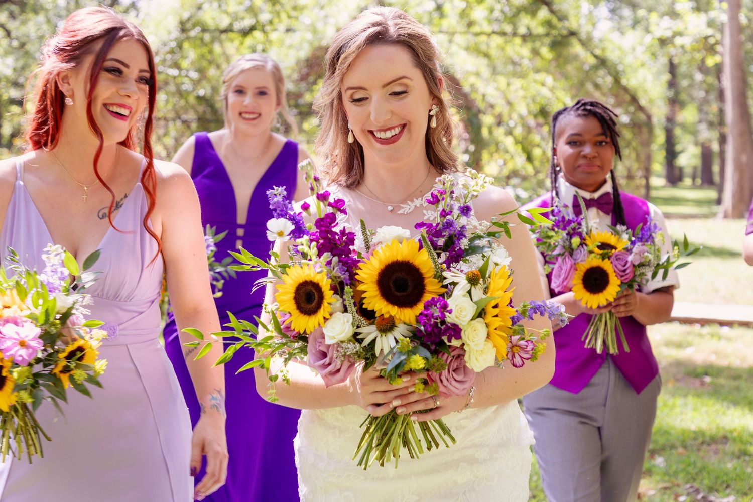 Wedding party in purple dresses holds sunflower bouquets outdoors.