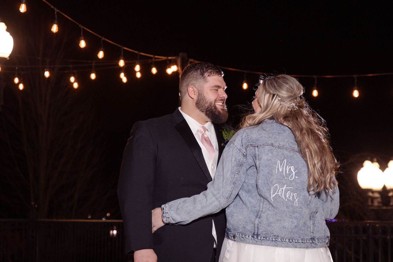 Romantic evening photos of newlyweds under string lights with bride wearing custom denim jacket.