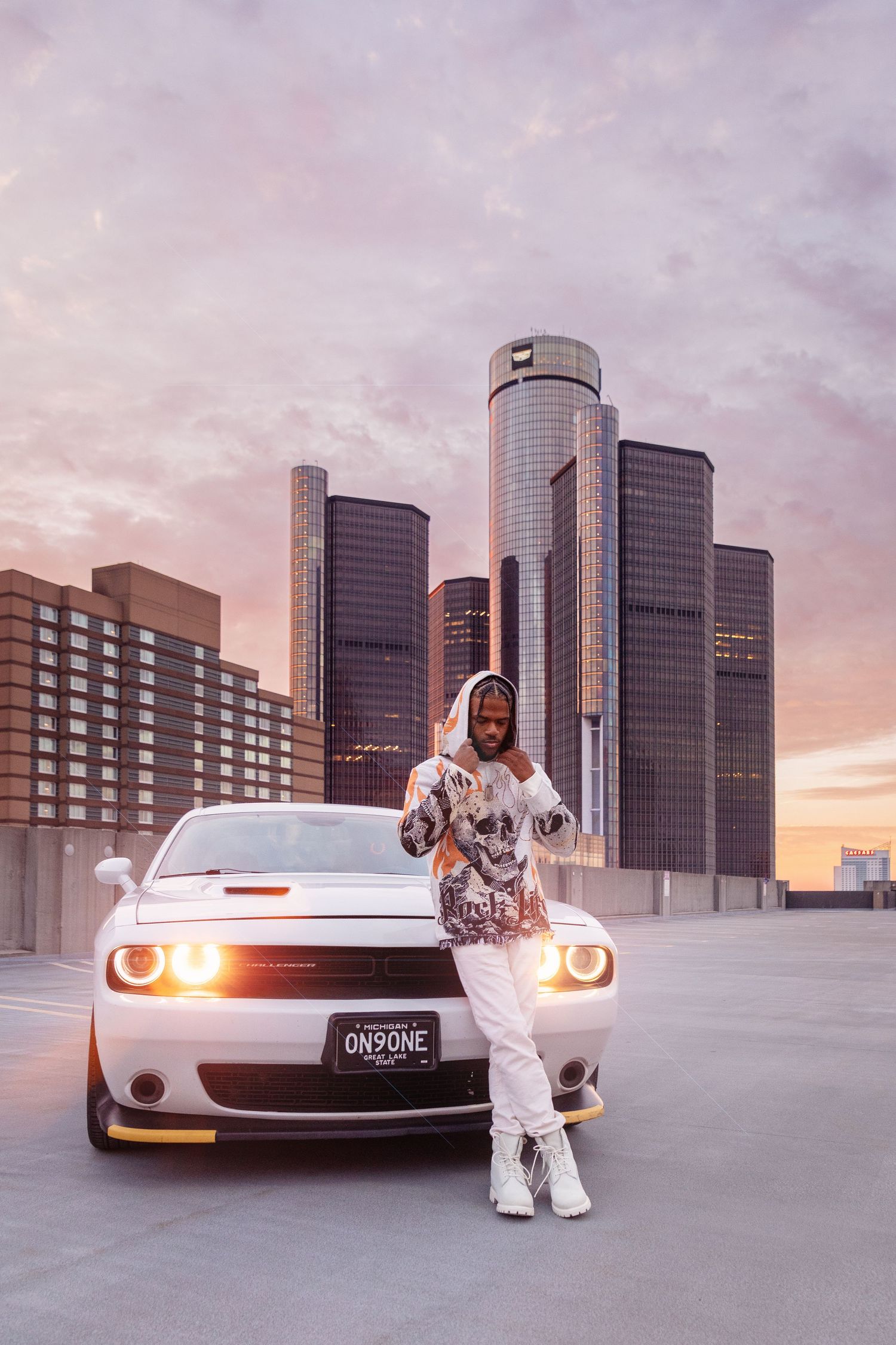 A white Dodge Challenger sits in front of Detroit's iconic Renaissance Center during golden hour.