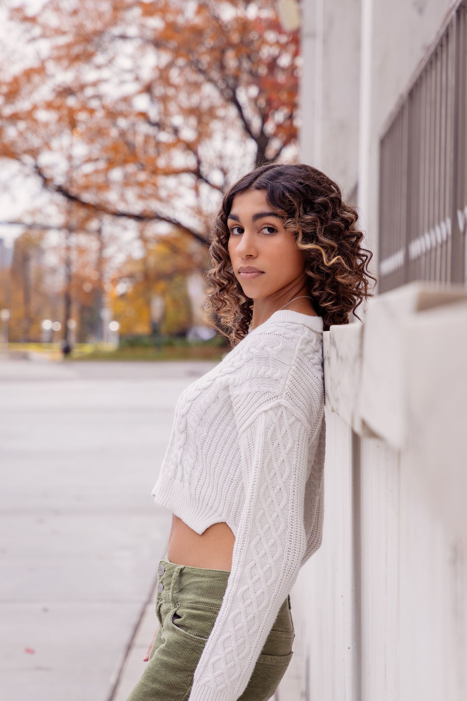 A fashion portrait showing a white cable knit sweater and curly hair against autumn trees.