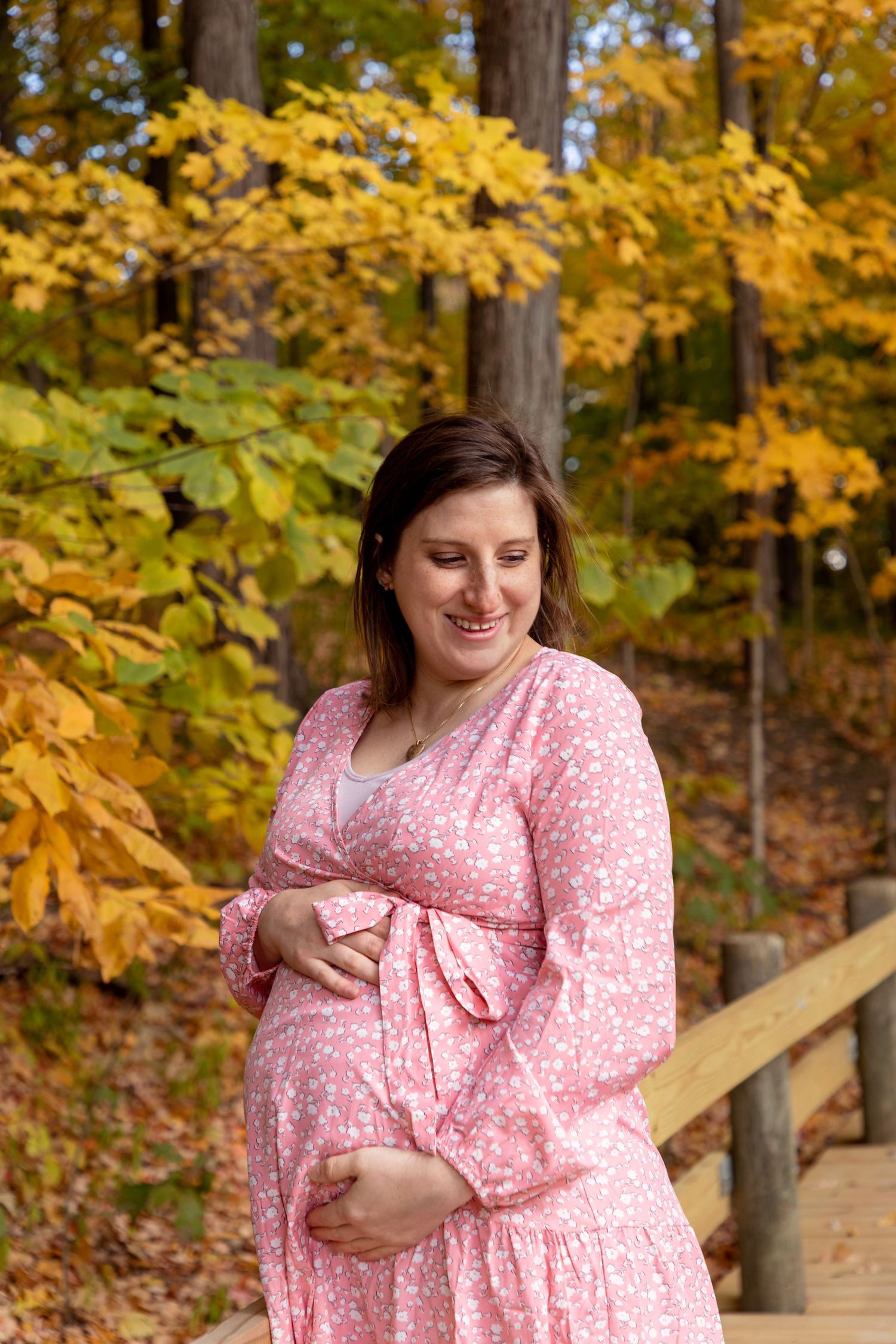 An expectant mother in a pink floral dress smiles while holding her belly amid fall foliage.