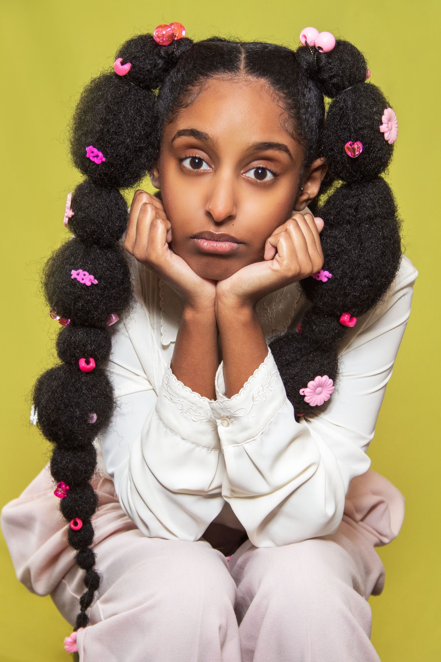 Young model in white outfit with pigtails decorated with pink flowers against yellow background.