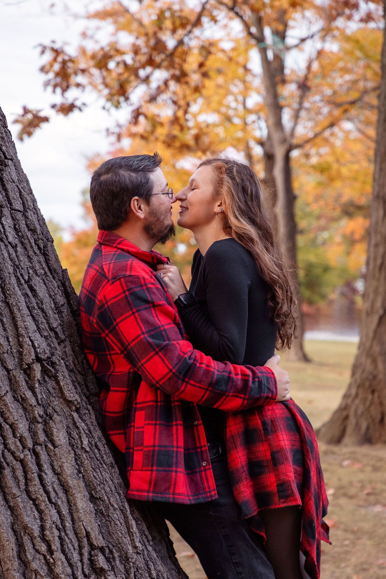 Couple embraces romantically against autumn trees wearing red plaid and black.