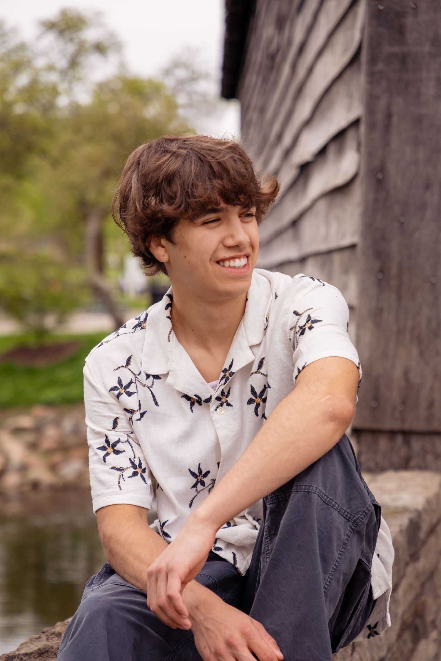 Young person in floral print shirt sitting casually outdoors against rustic wooden backdrop.