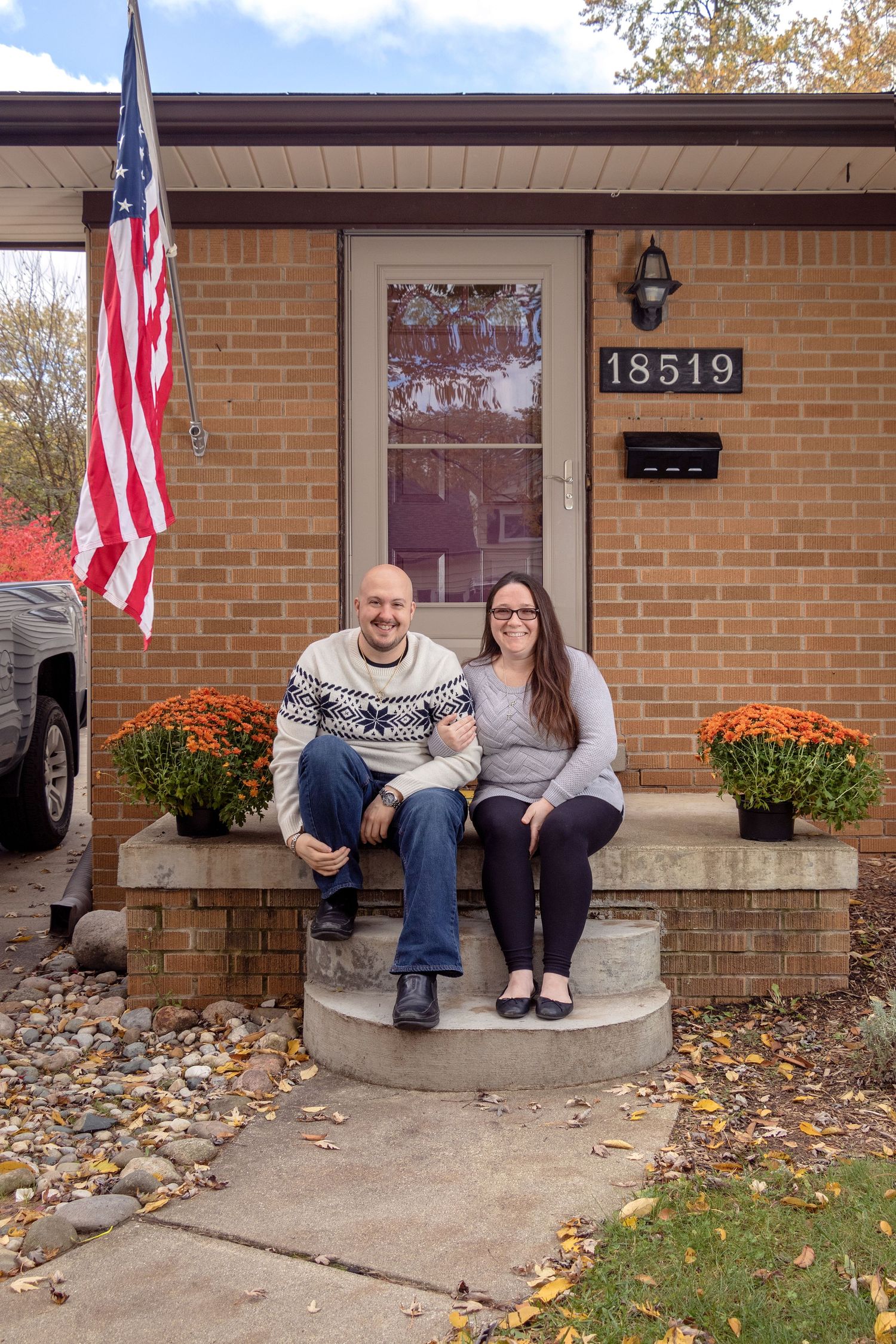 Couple sitting on front porch steps of brick house with American flag and fall decorations.