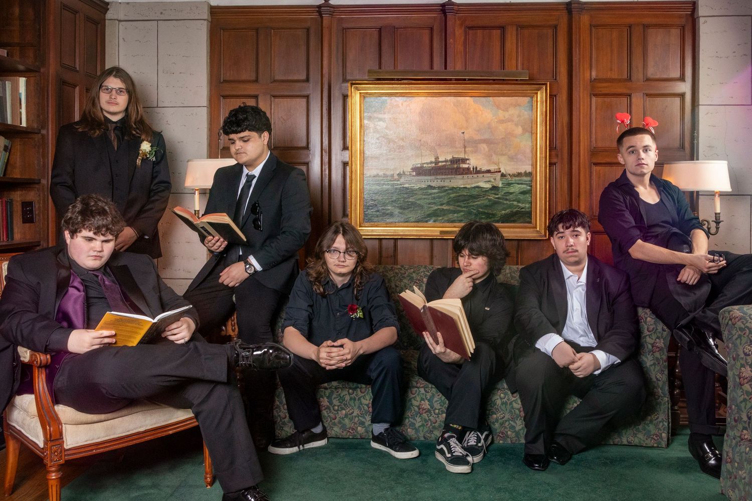 Group of formally dressed people sitting on green couch in front of wood-paneled wall with artwork.