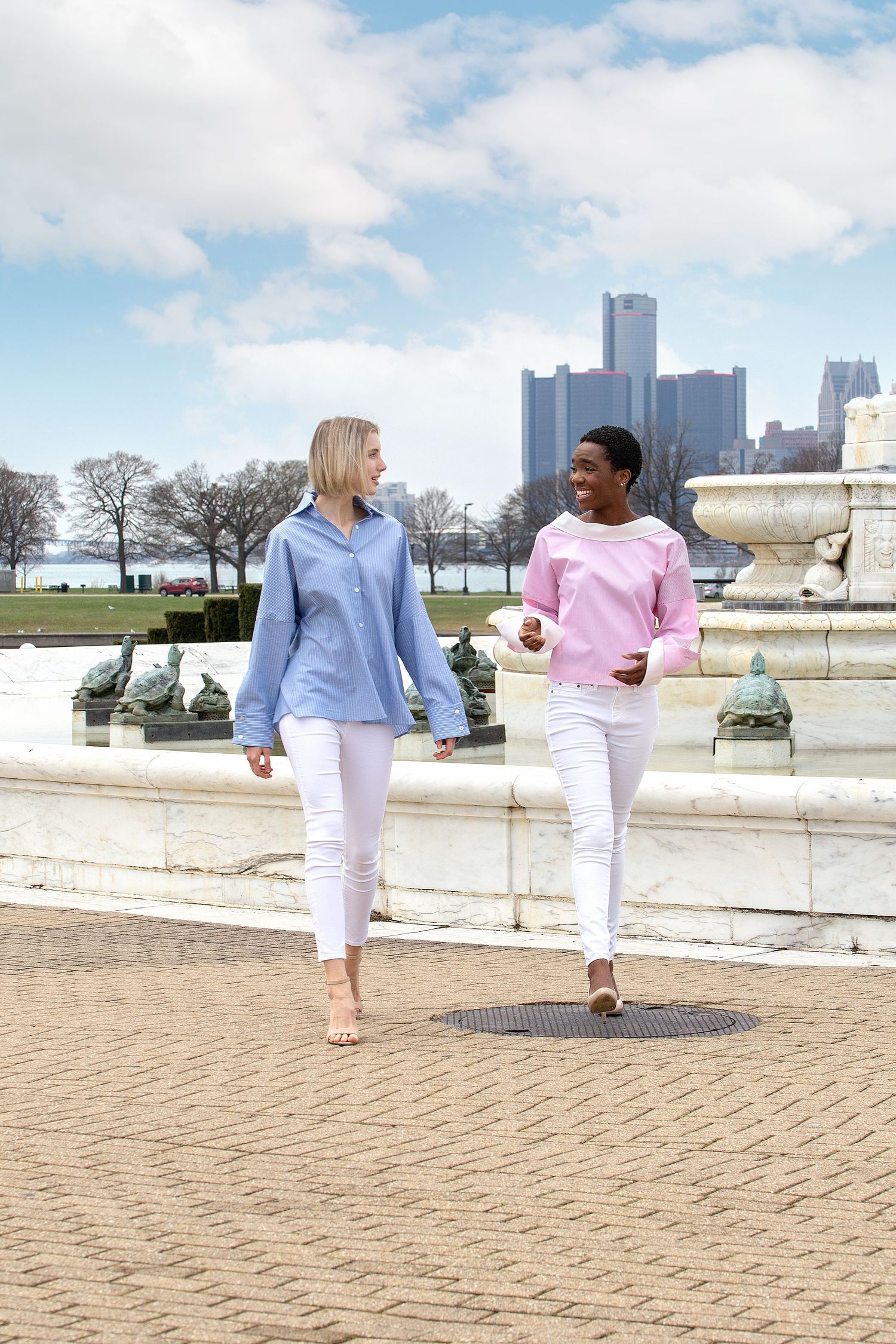 People walking along Detroit riverfront on sunny day with city skyline in background.