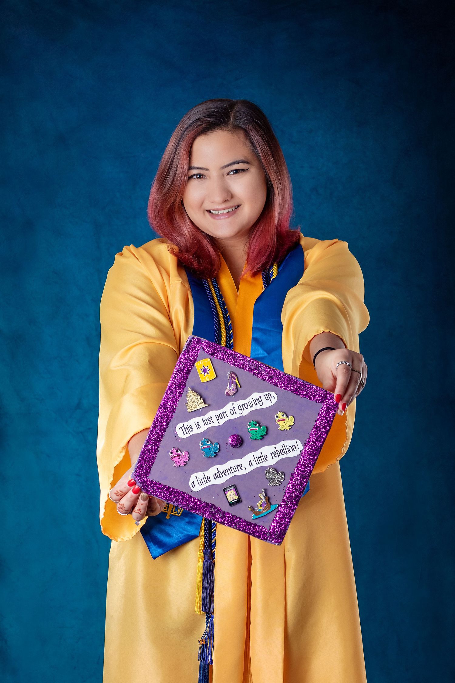 Person in yellow graduation gown holds decorated graduation cap against blue background.