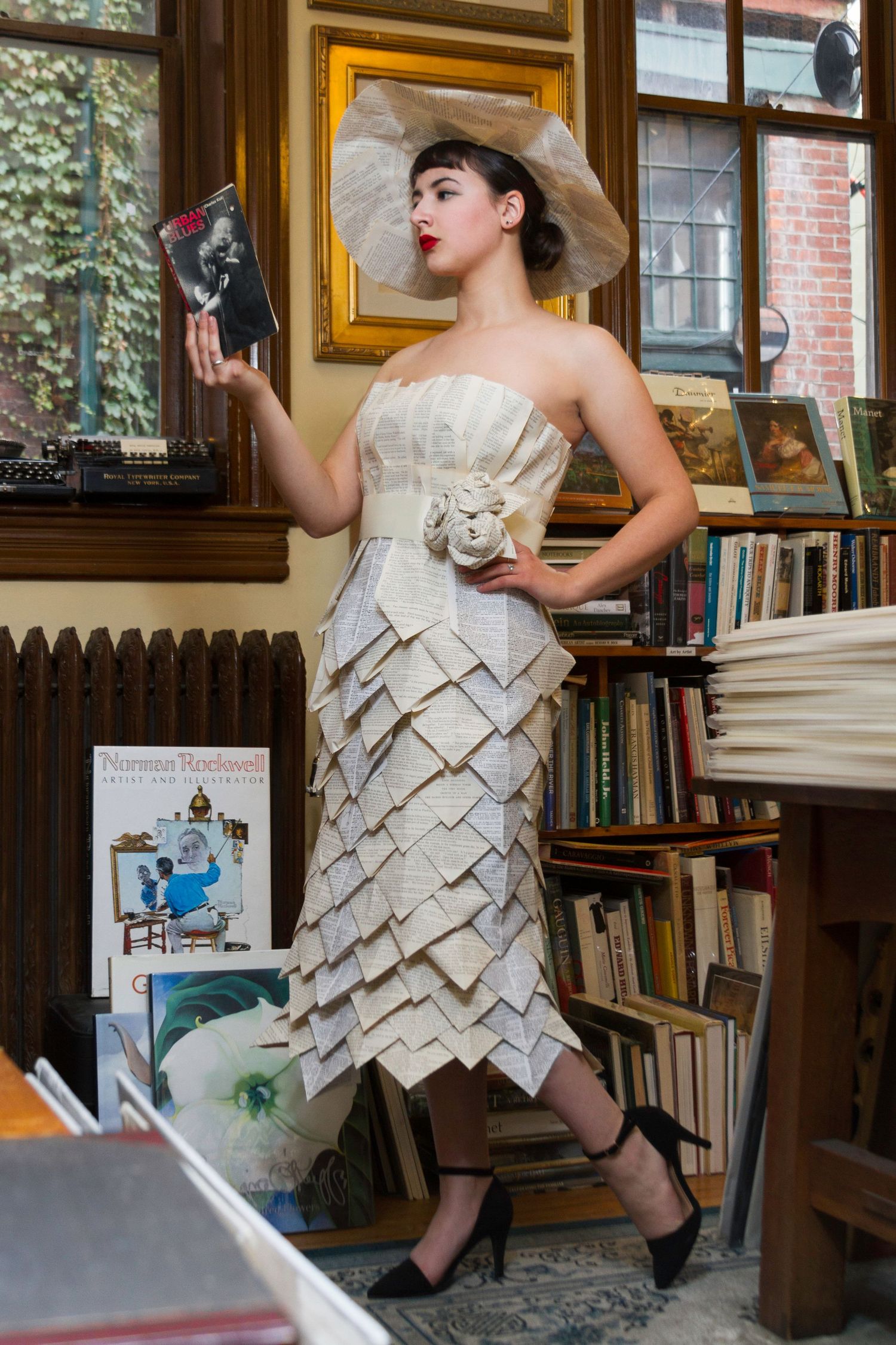 Model in paper dress made from book pages poses in library setting.