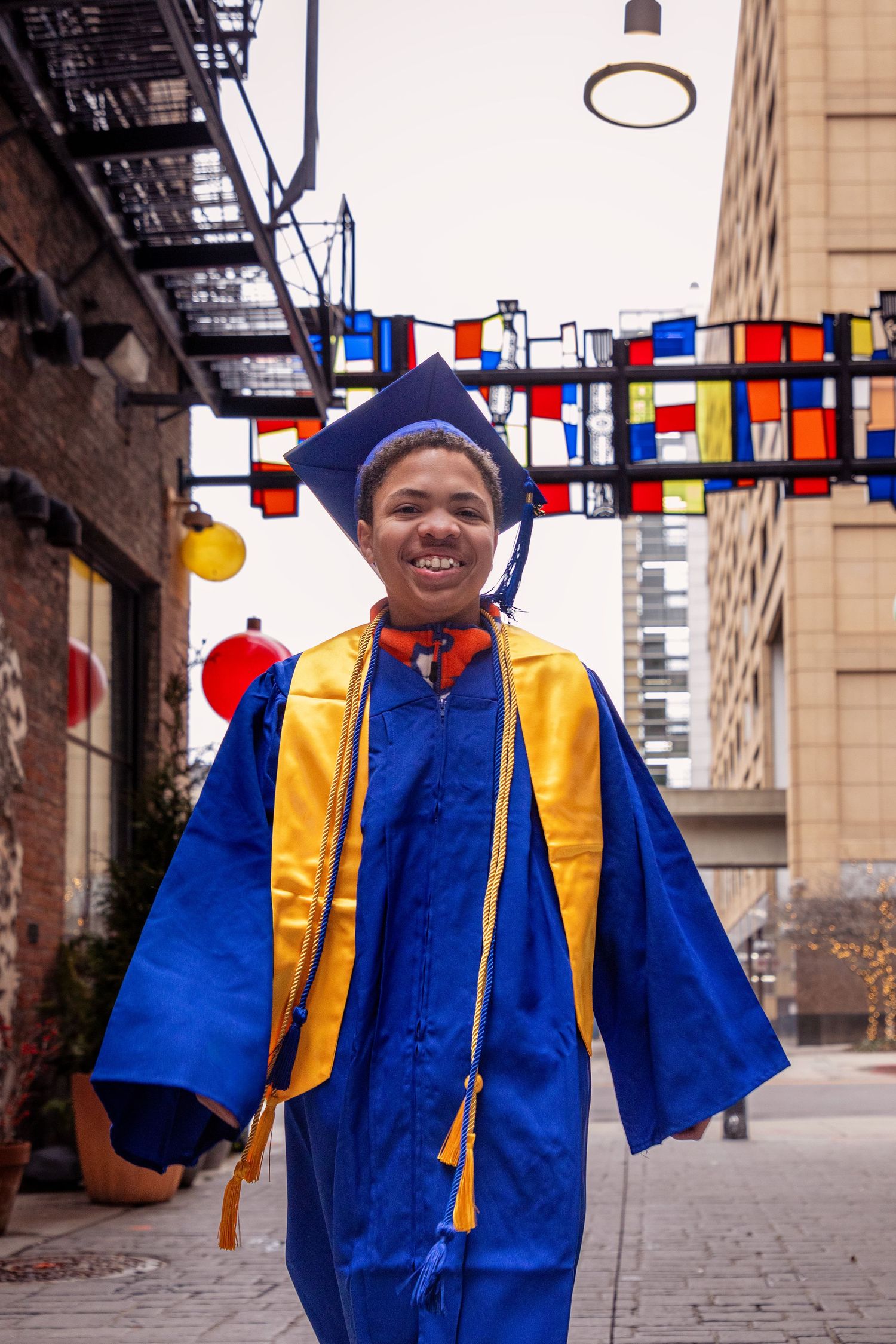 Person in bright blue and gold graduation attire walking outdoors with colorful banners overhead.