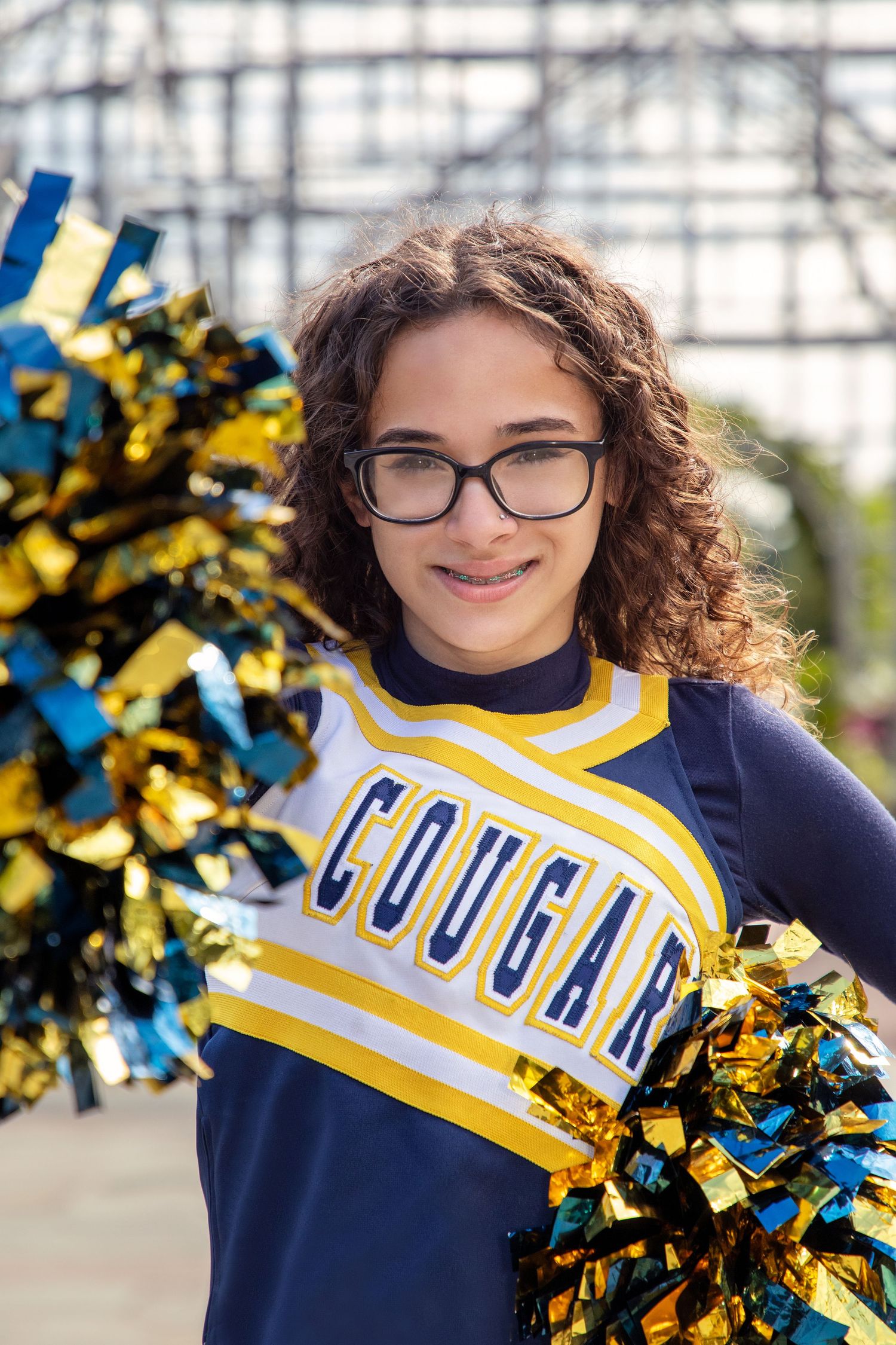 Cheerleader in navy and yellow uniform poses with pom poms and bright smile.