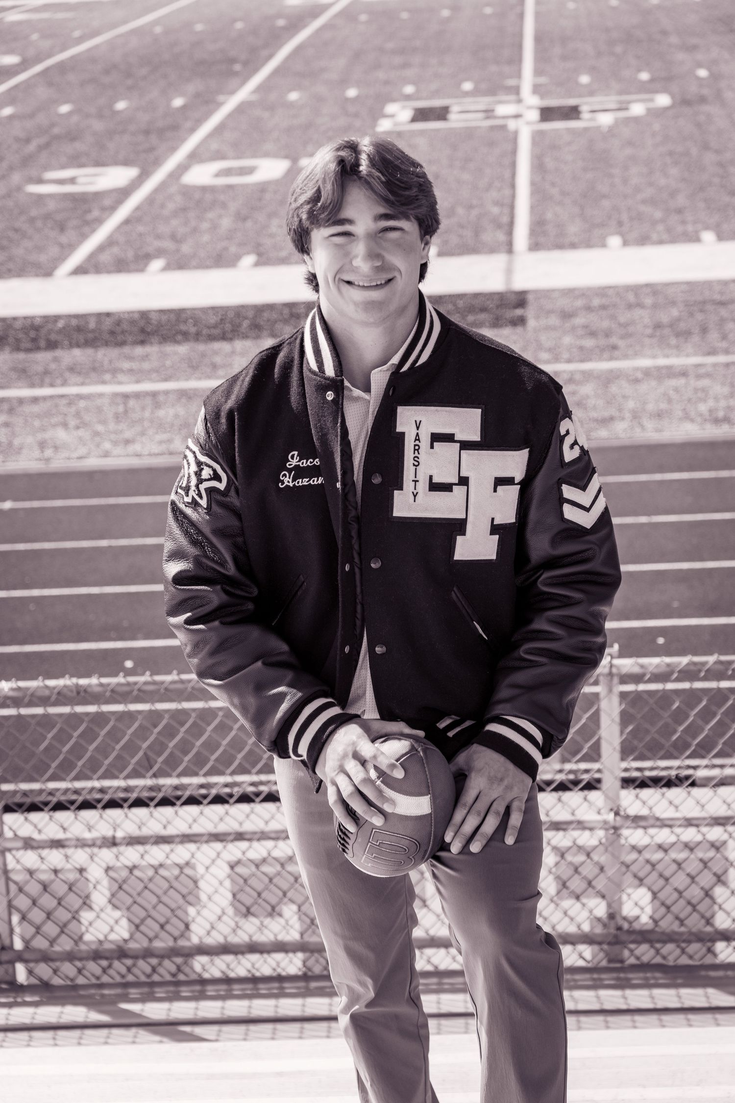 Black and white photo of student in varsity jacket sitting in stadium bleachers.