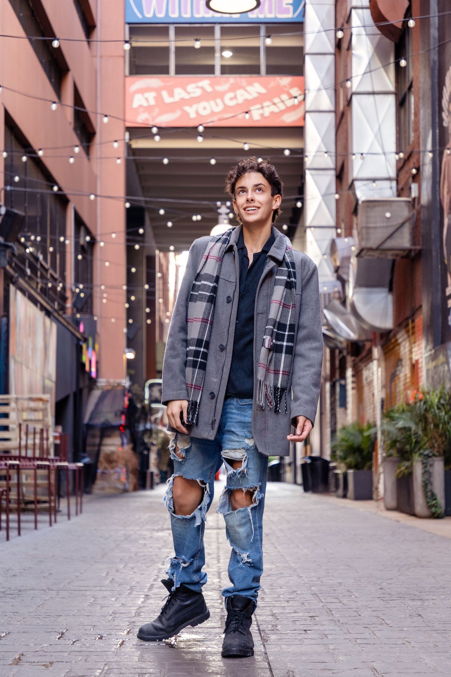 Person in distressed jeans and jacket poses along urban storefront walkway.