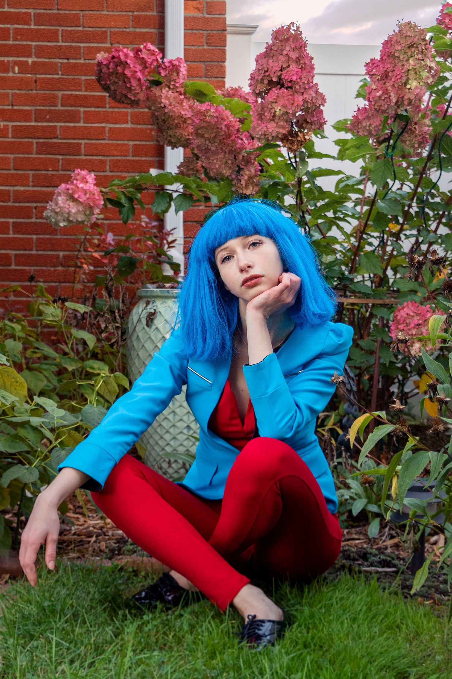 Figure in bright blue jacket and red pants poses casually on grass near flowering bushes and brick wall.