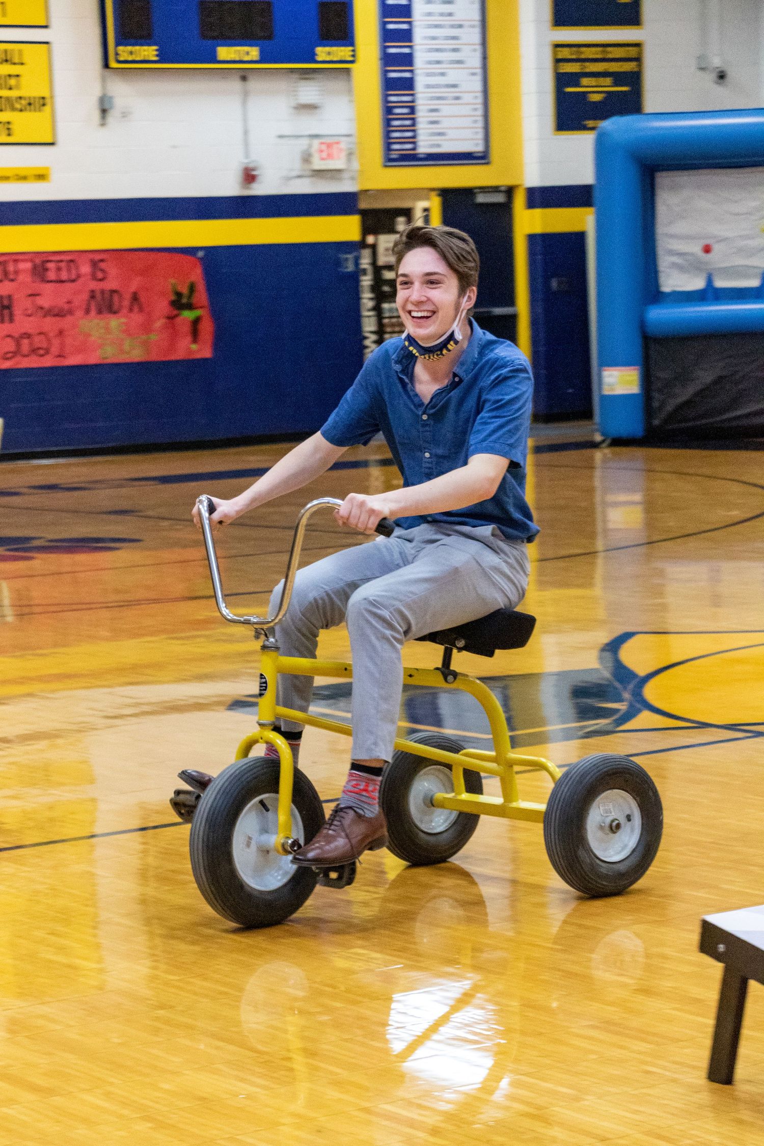 Someone rides a yellow tricycle across a shiny gymnasium floor with blue and yellow decor.