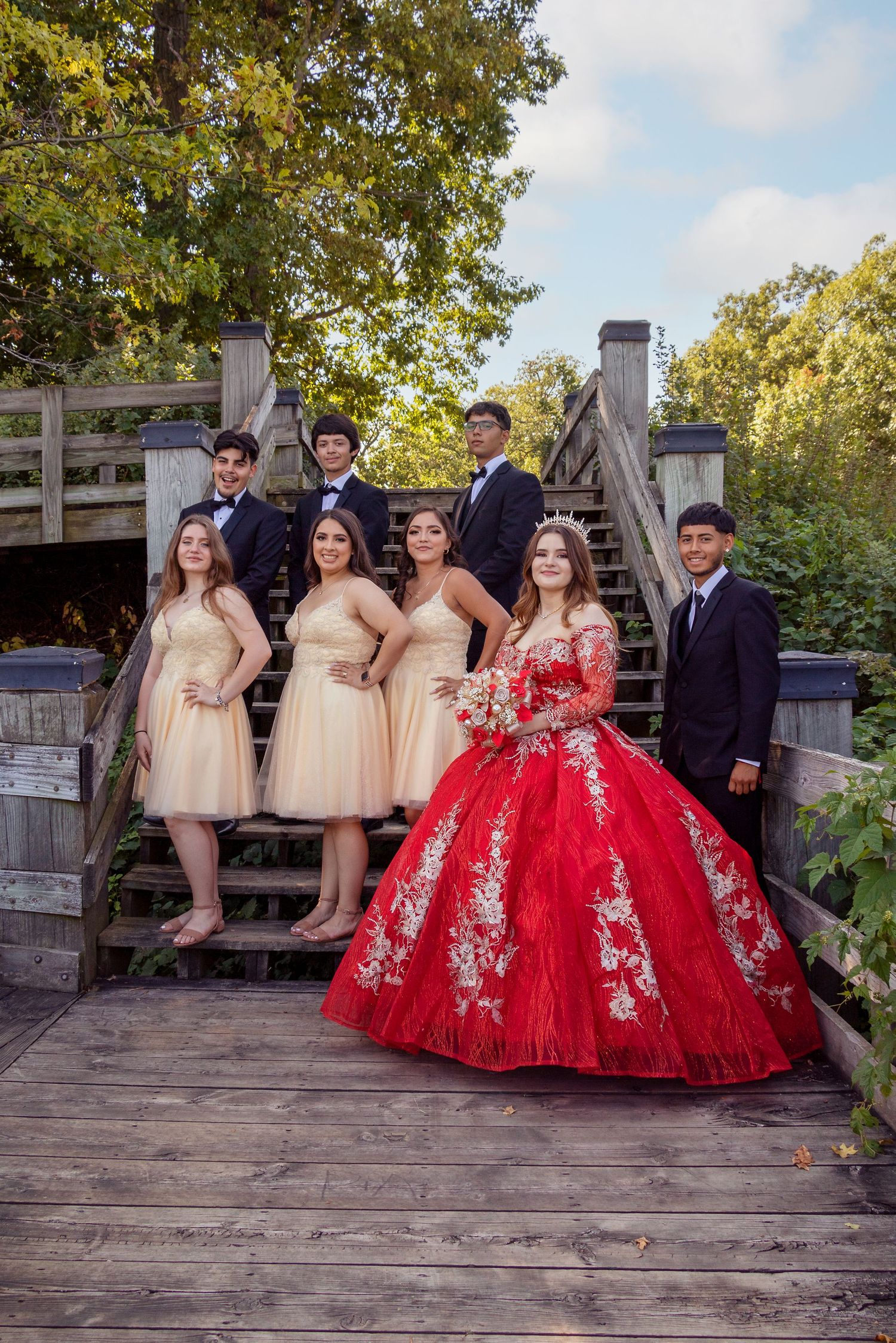 A quinceañera court posing on wooden deck wearing red and champagne formal attire.