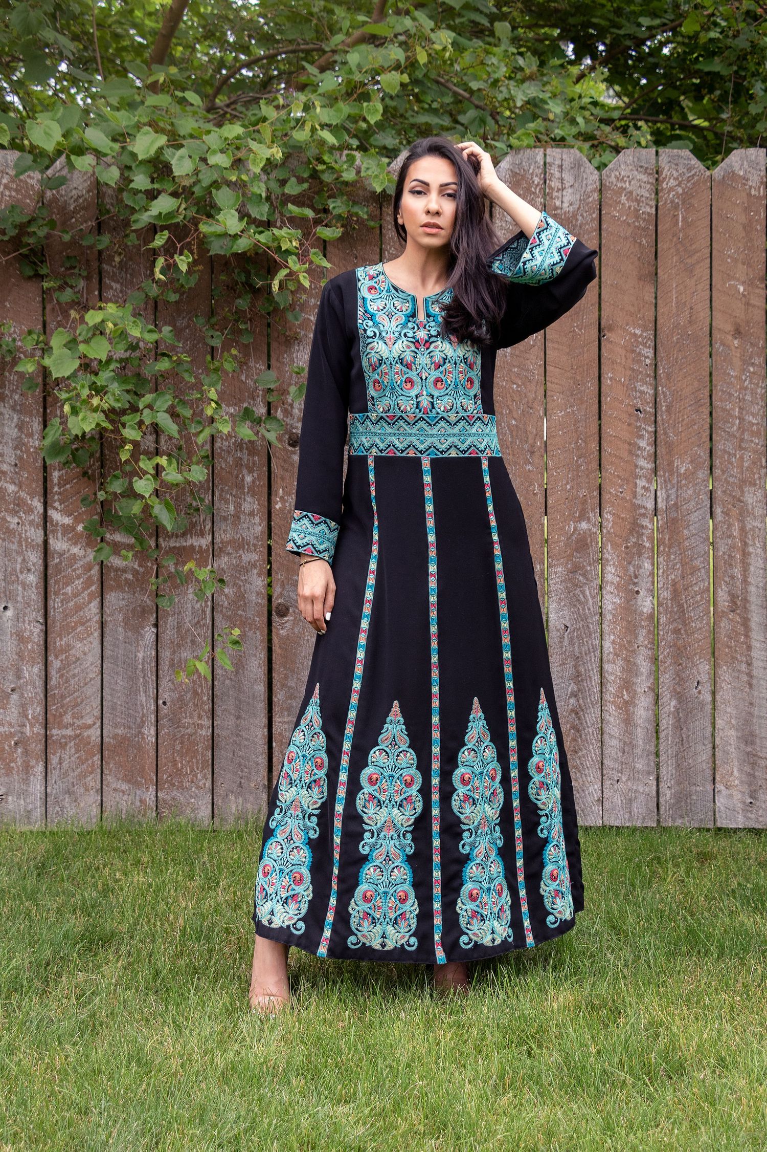 Long black dress with turquoise embroidered details shown against wooden fence backdrop.