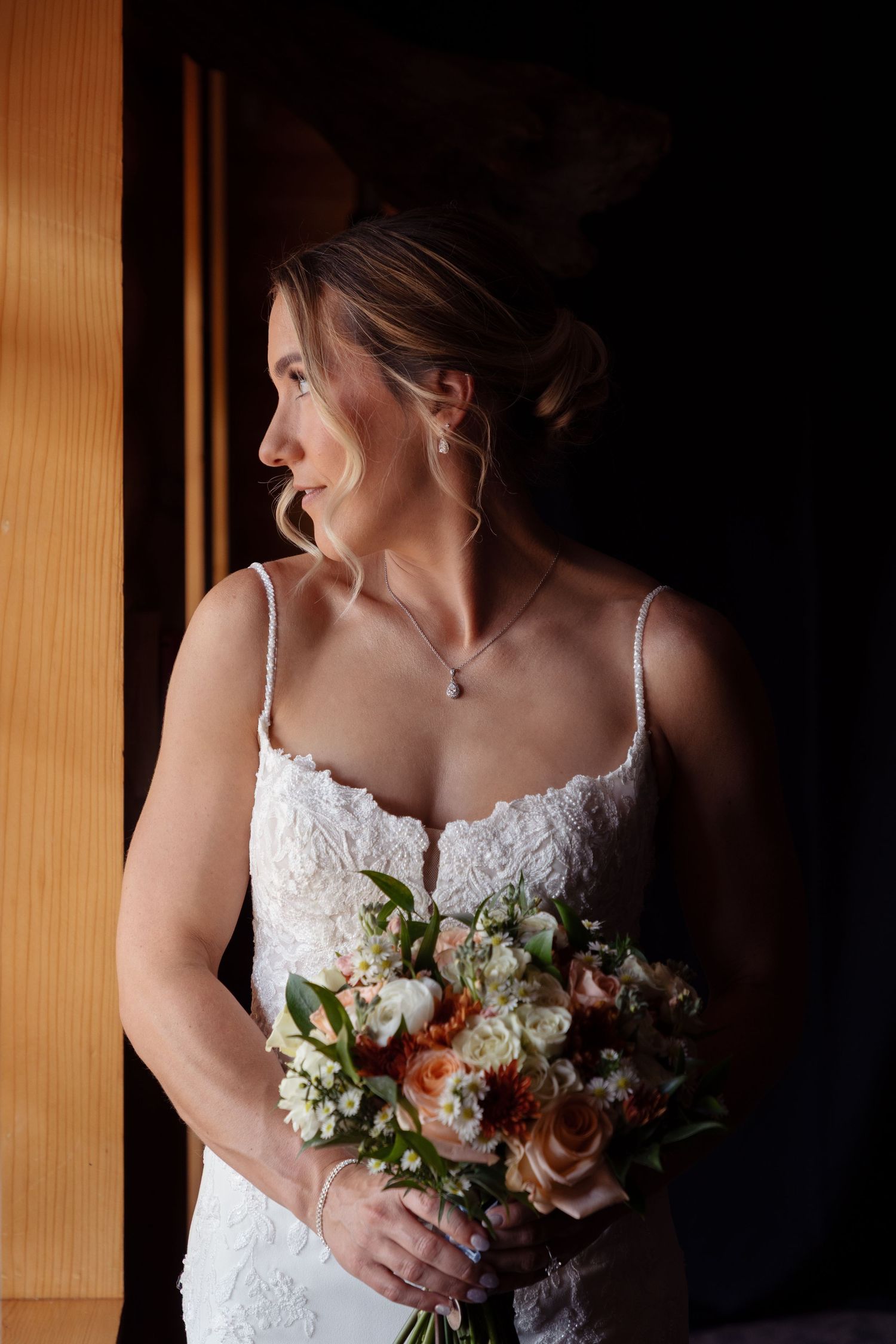 Dramatic window light illuminates bride holding delicate floral bouquet in white lace dress.