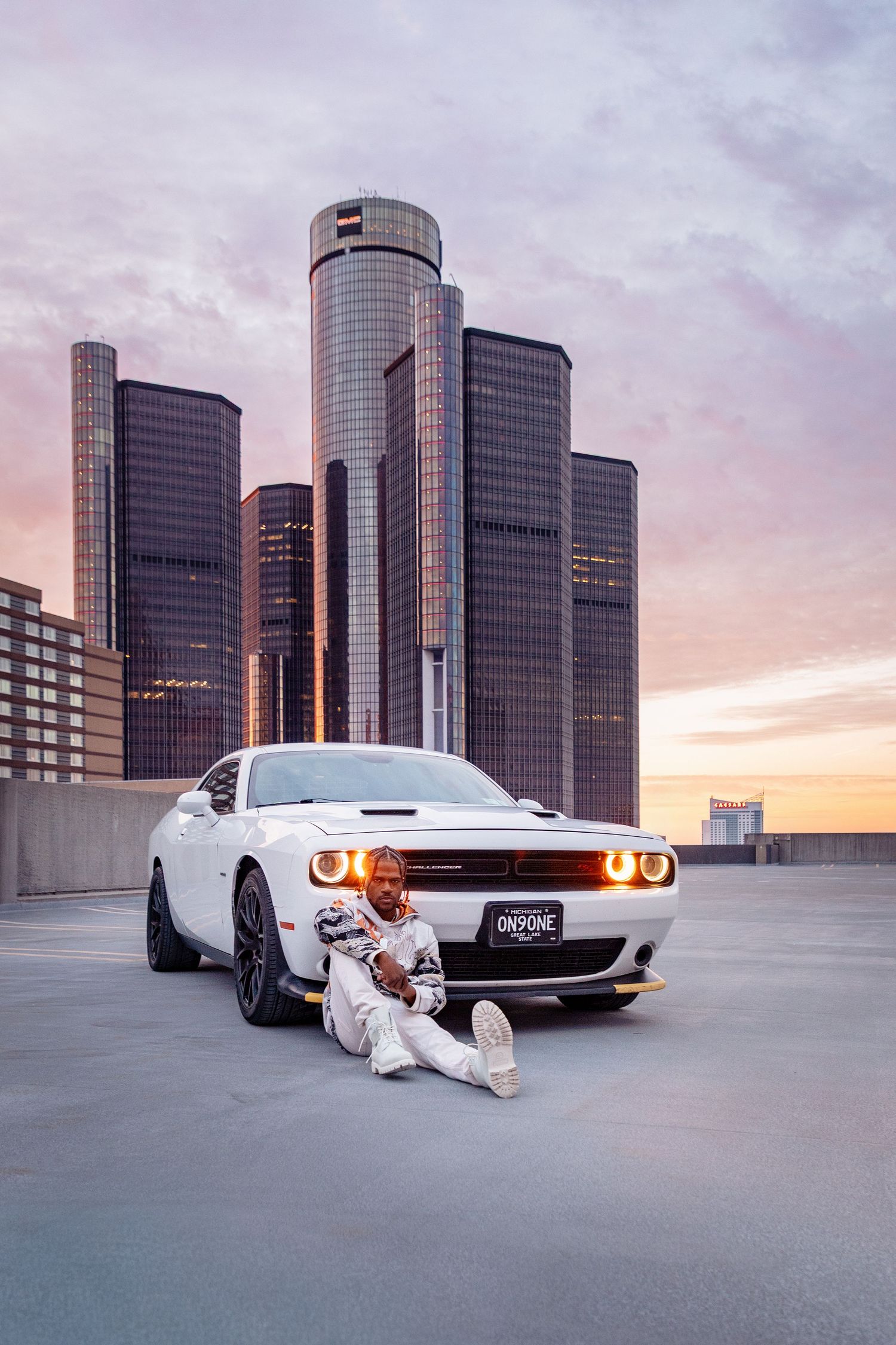 A white Dodge Challenger parked in front of Detroit's Renaissance Center at sunset.