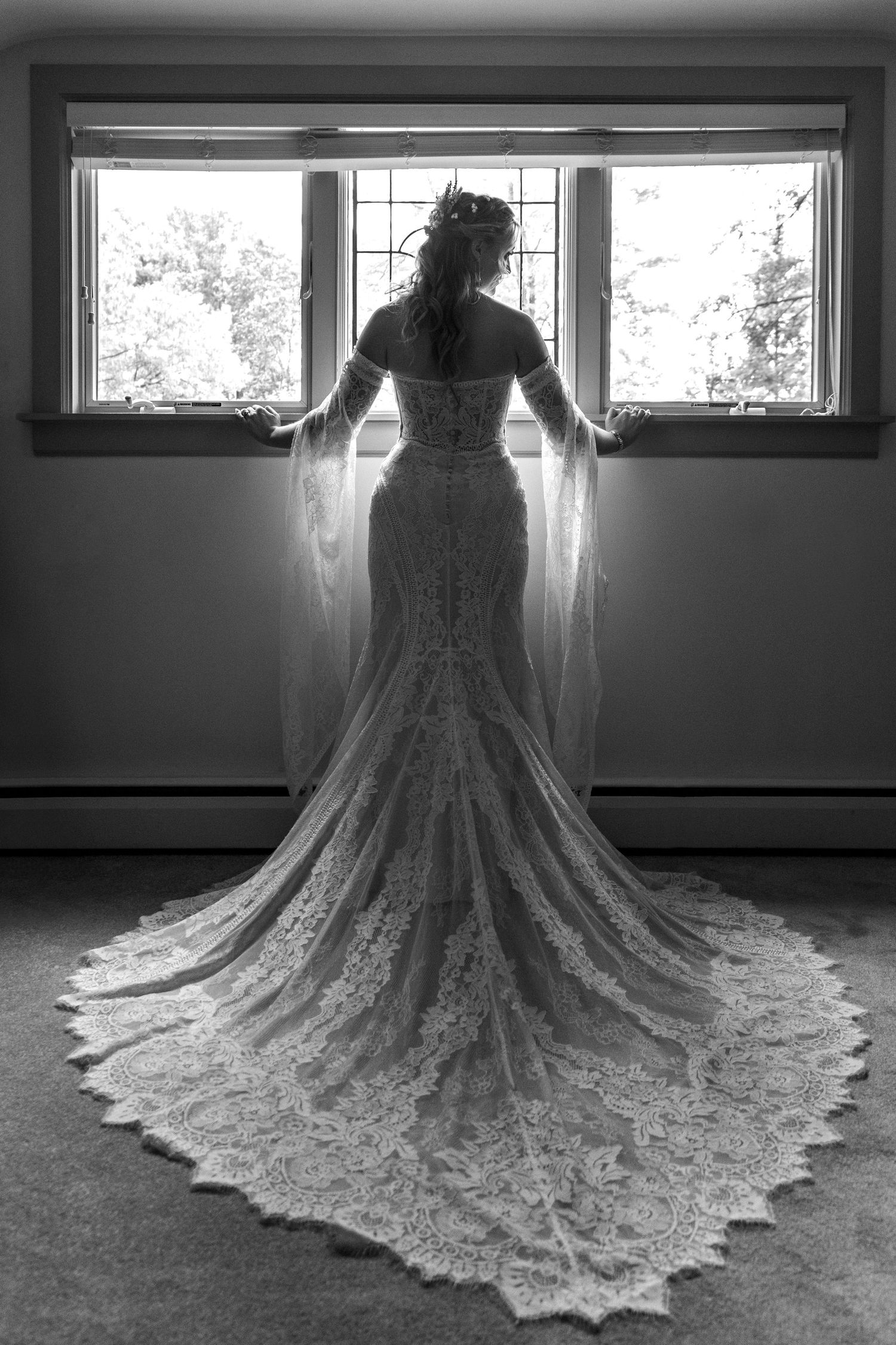 Black and white photograph of wedding dress train with intricate lace details near windows.