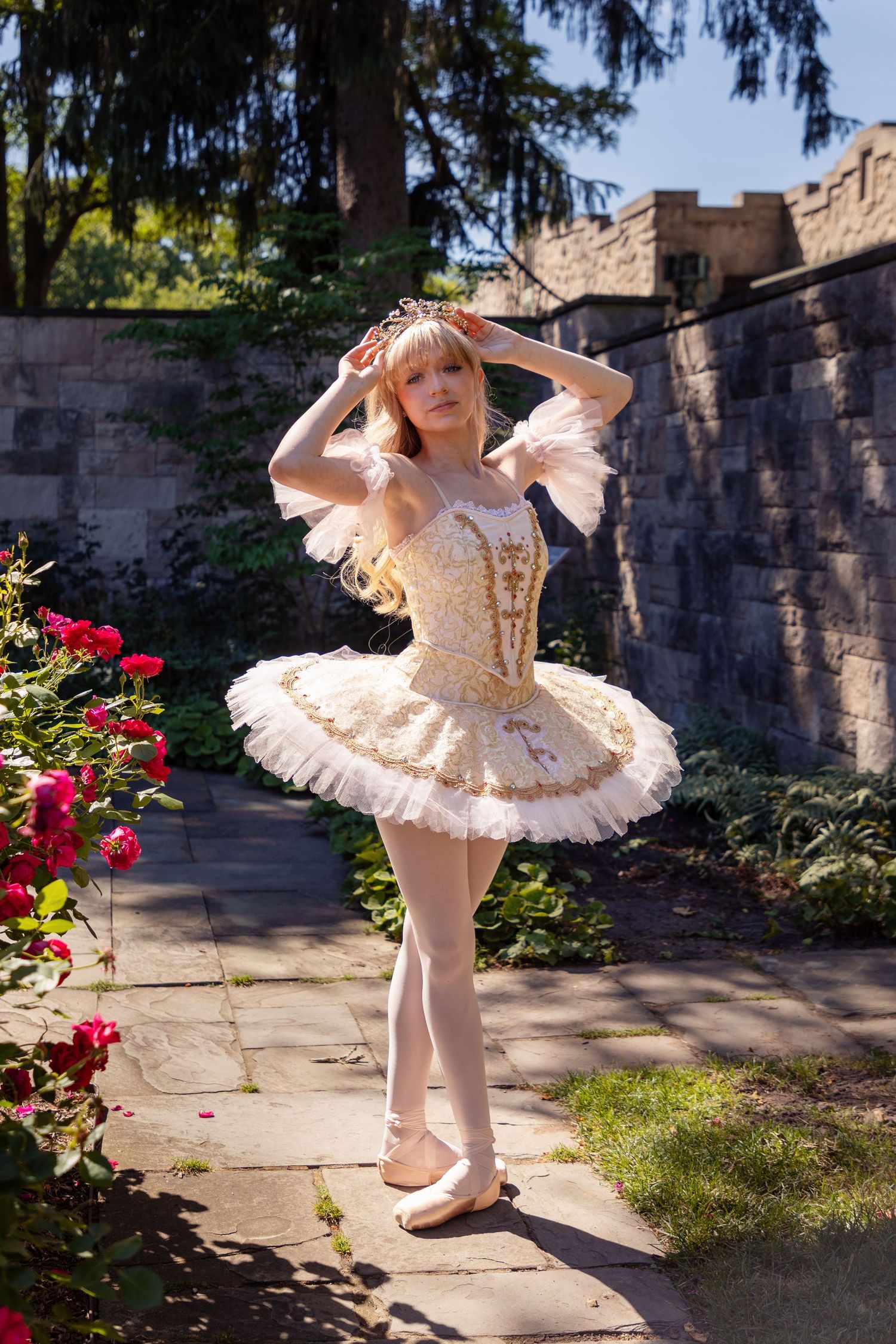 A ballet dancer in a white tutu poses gracefully in a garden setting with stone walls and pink flowers.