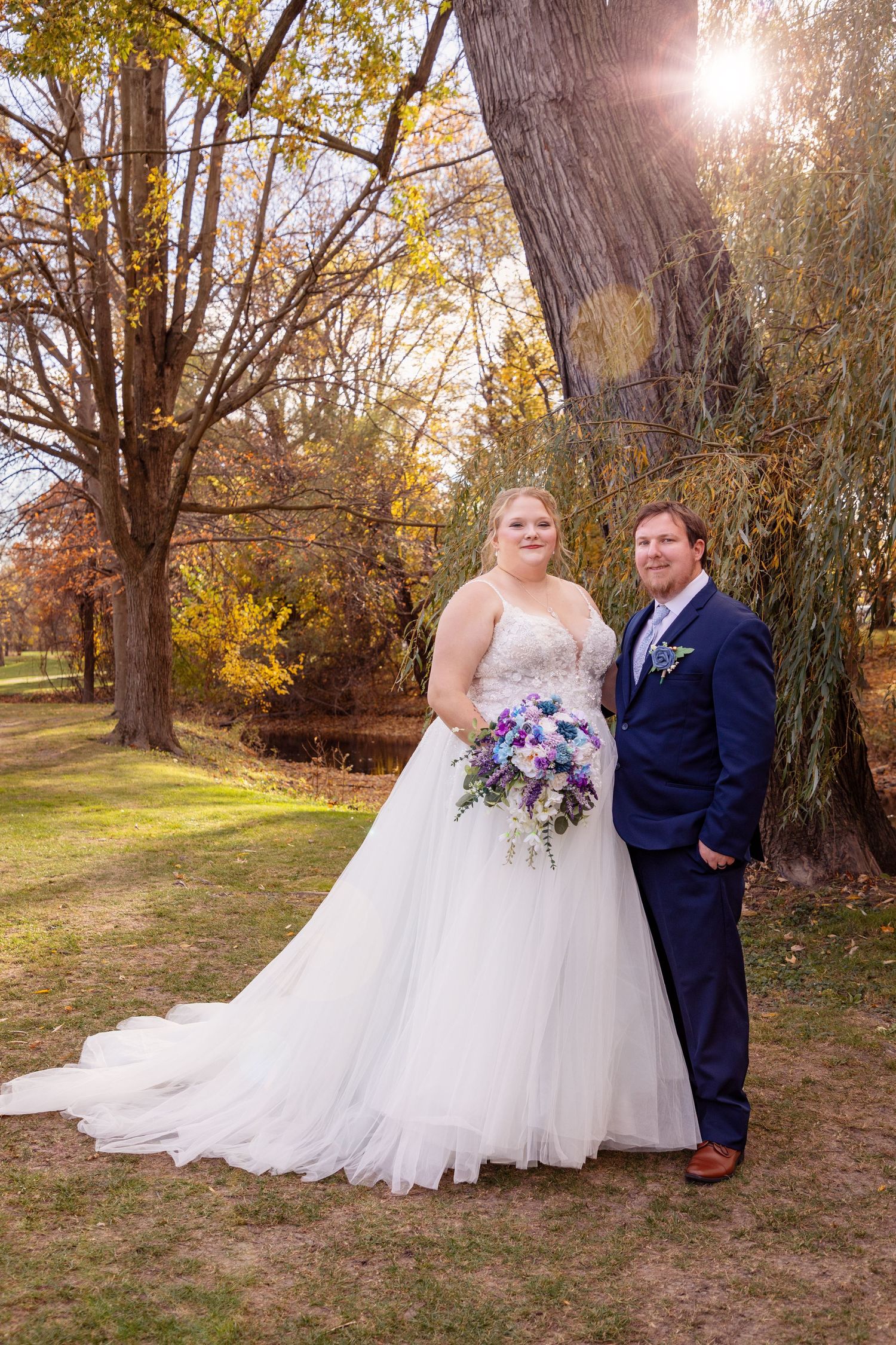 Newlywed couple poses together in a scenic autumn park setting with golden leaves and sunlight.
