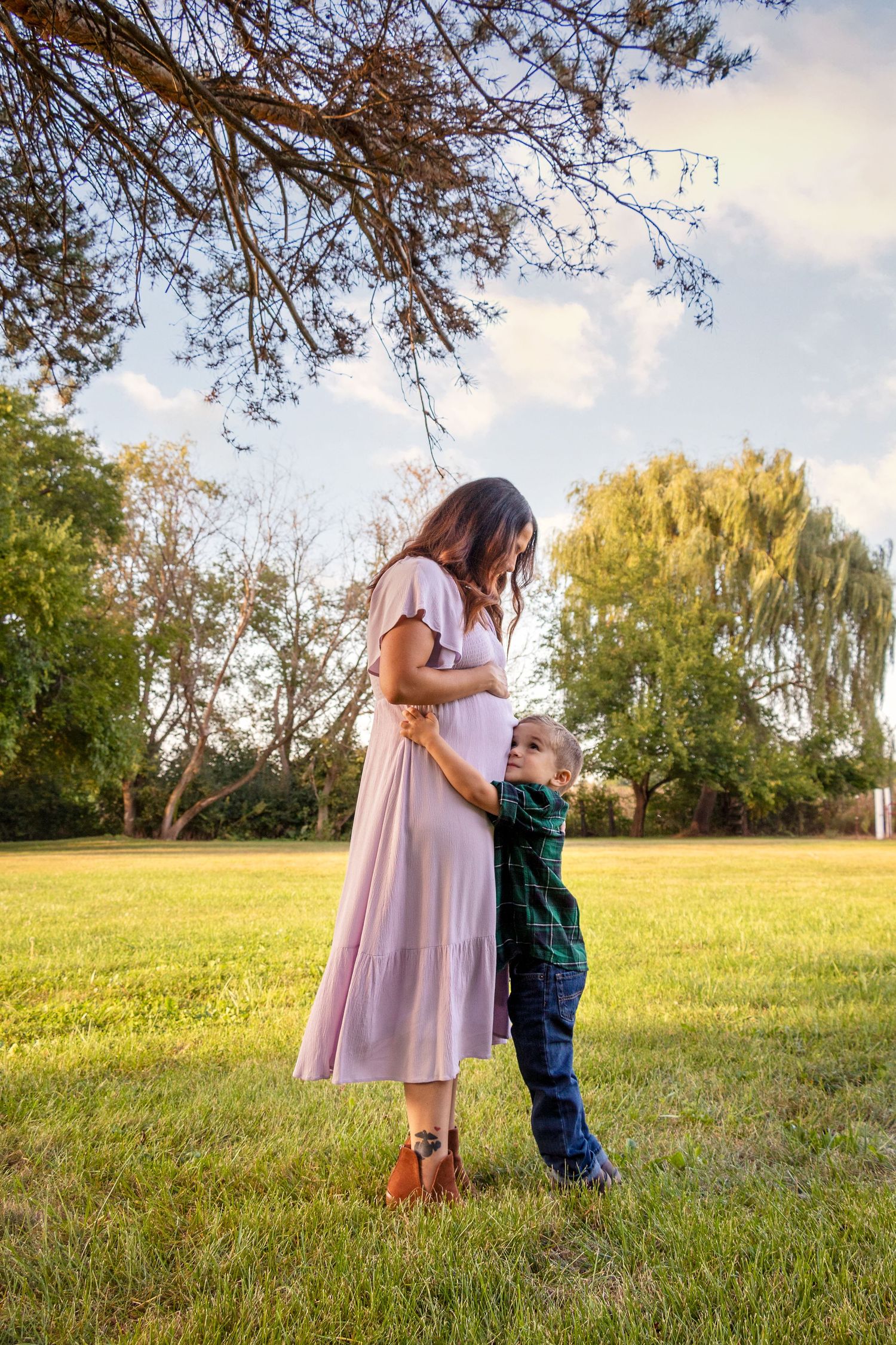 A tender moment captured in a sunlit field as a parent and child embrace beneath overhanging tree branches.