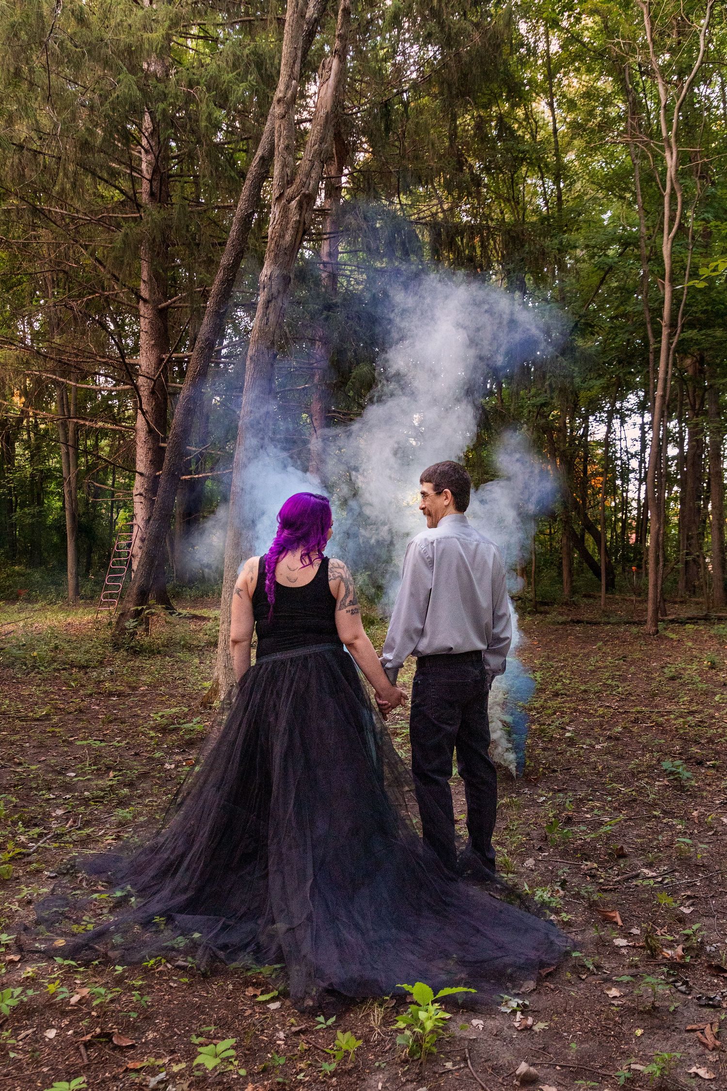 A couple stands in a misty forest with smoke effects, wearing formal attire including a dramatic black tulle gown.