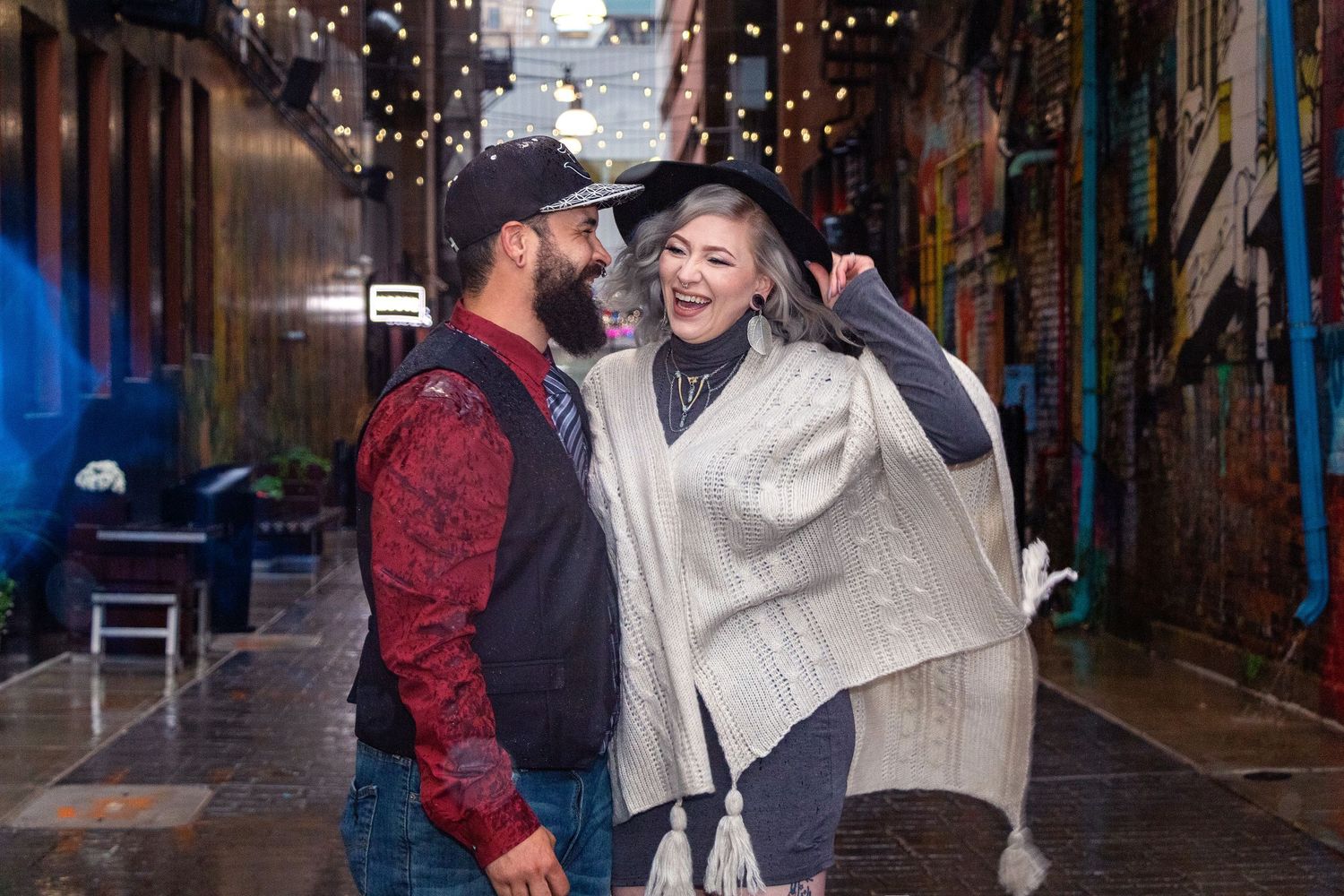 A couple shares a laugh together in an alley decorated with string lights.