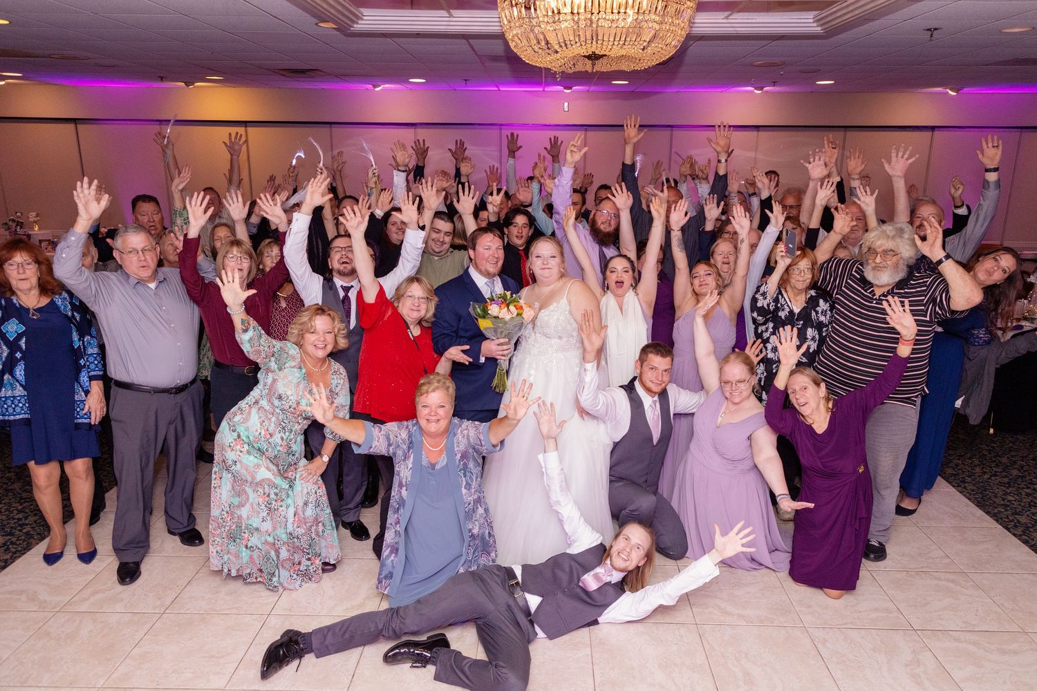 Large wedding reception crowd dancing and celebrating in an elegant ballroom with purple lighting.