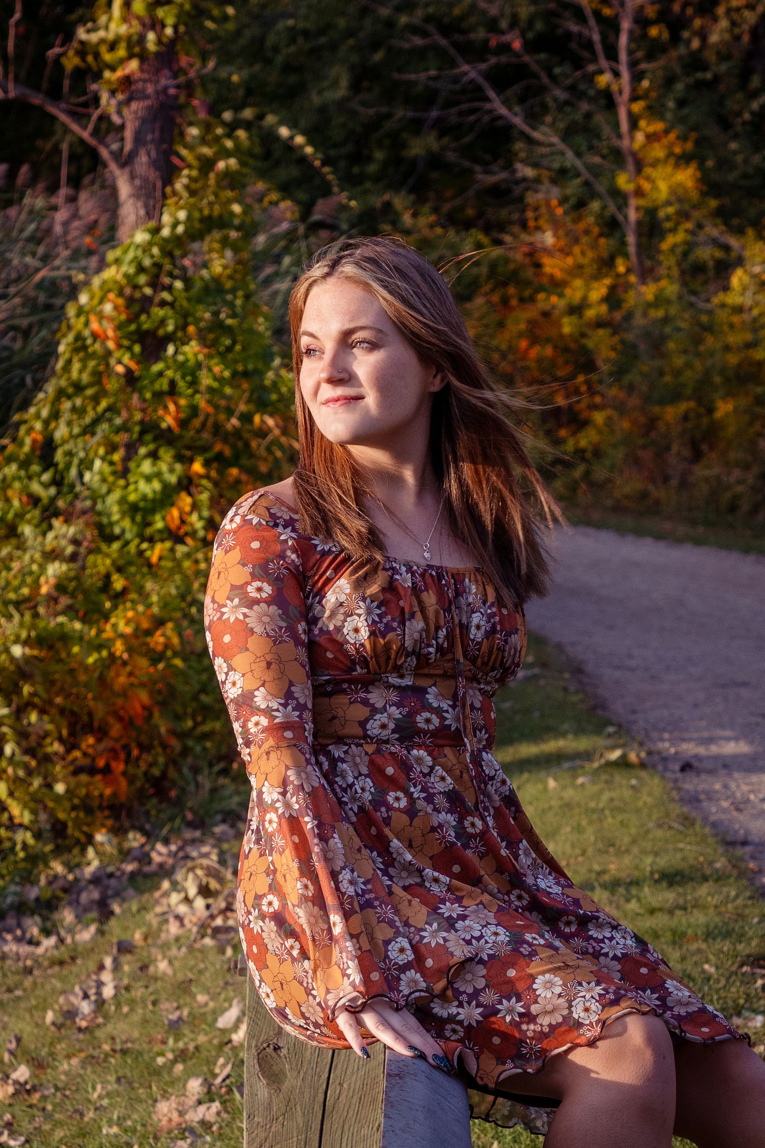 Person in floral dress poses on wooden bench during golden hour with autumn foliage background.