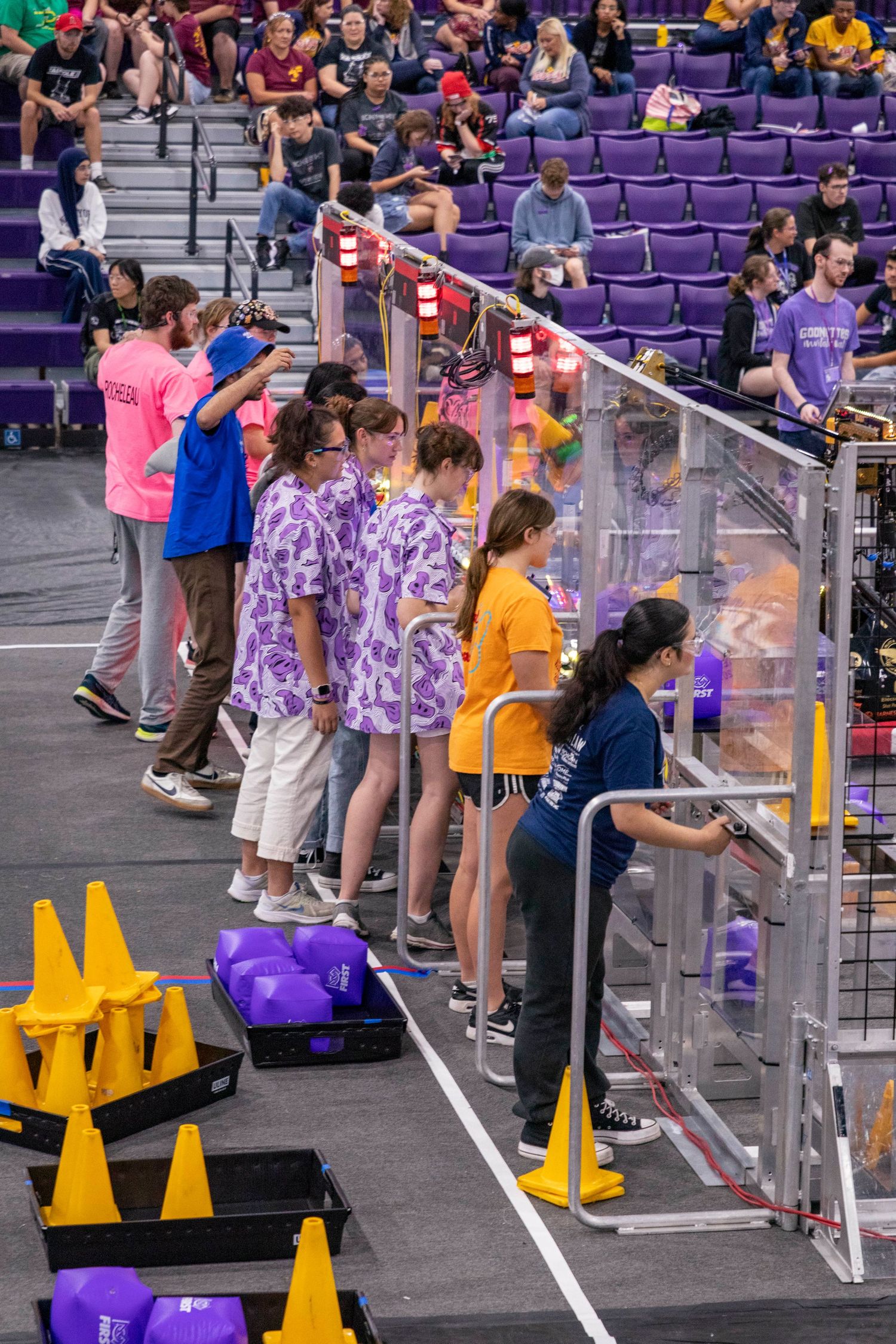 Students lined up along barriers in a gymnasium with purple seats.
