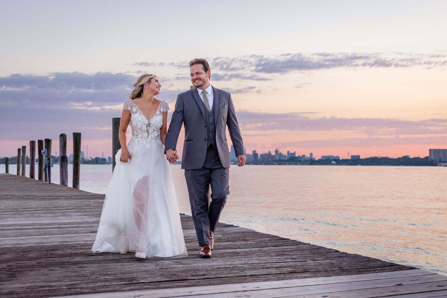 Couple walks along wooden pier at sunset with city skyline in background.