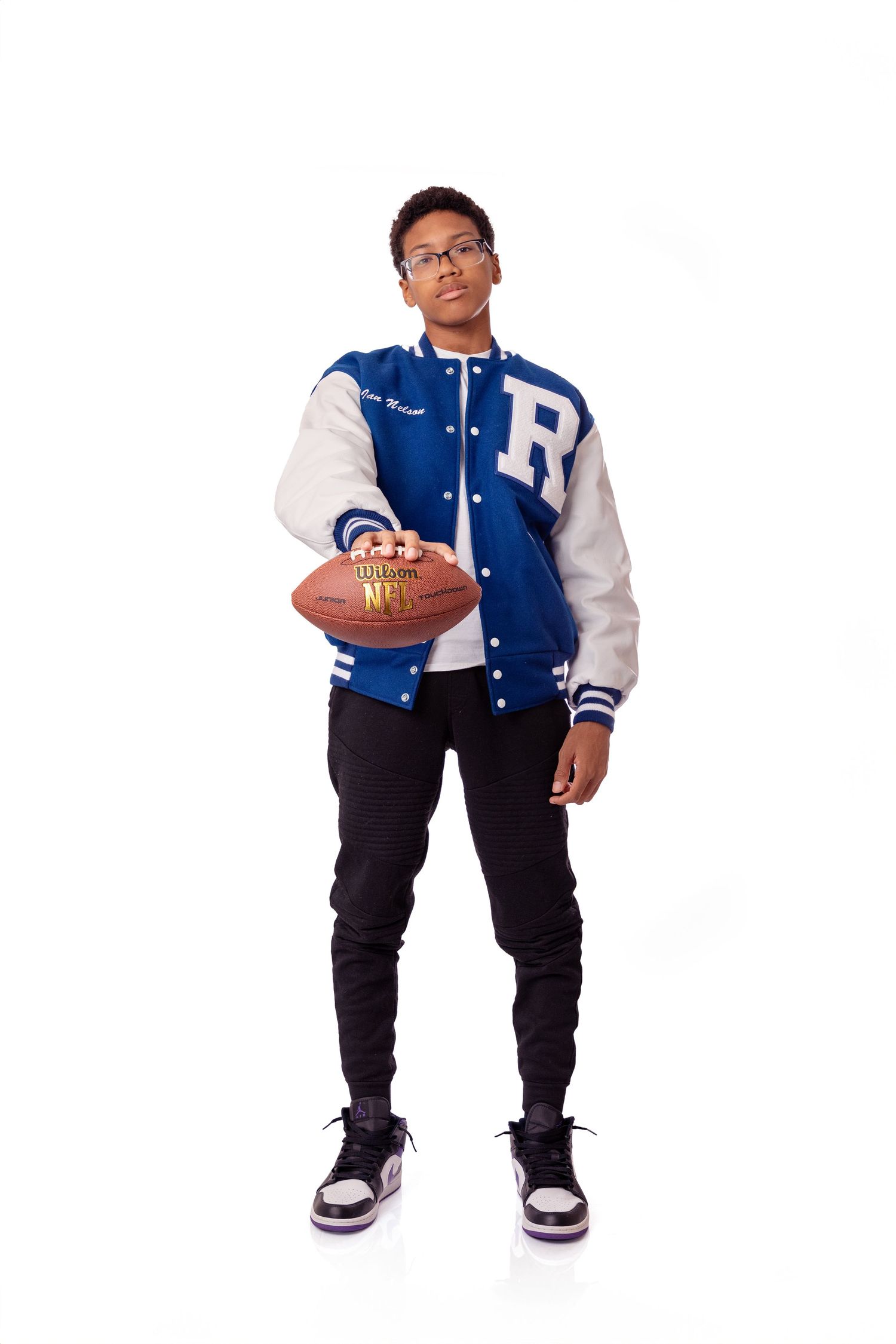 Young athlete poses with football wearing blue and white varsity jacket against white backdrop.