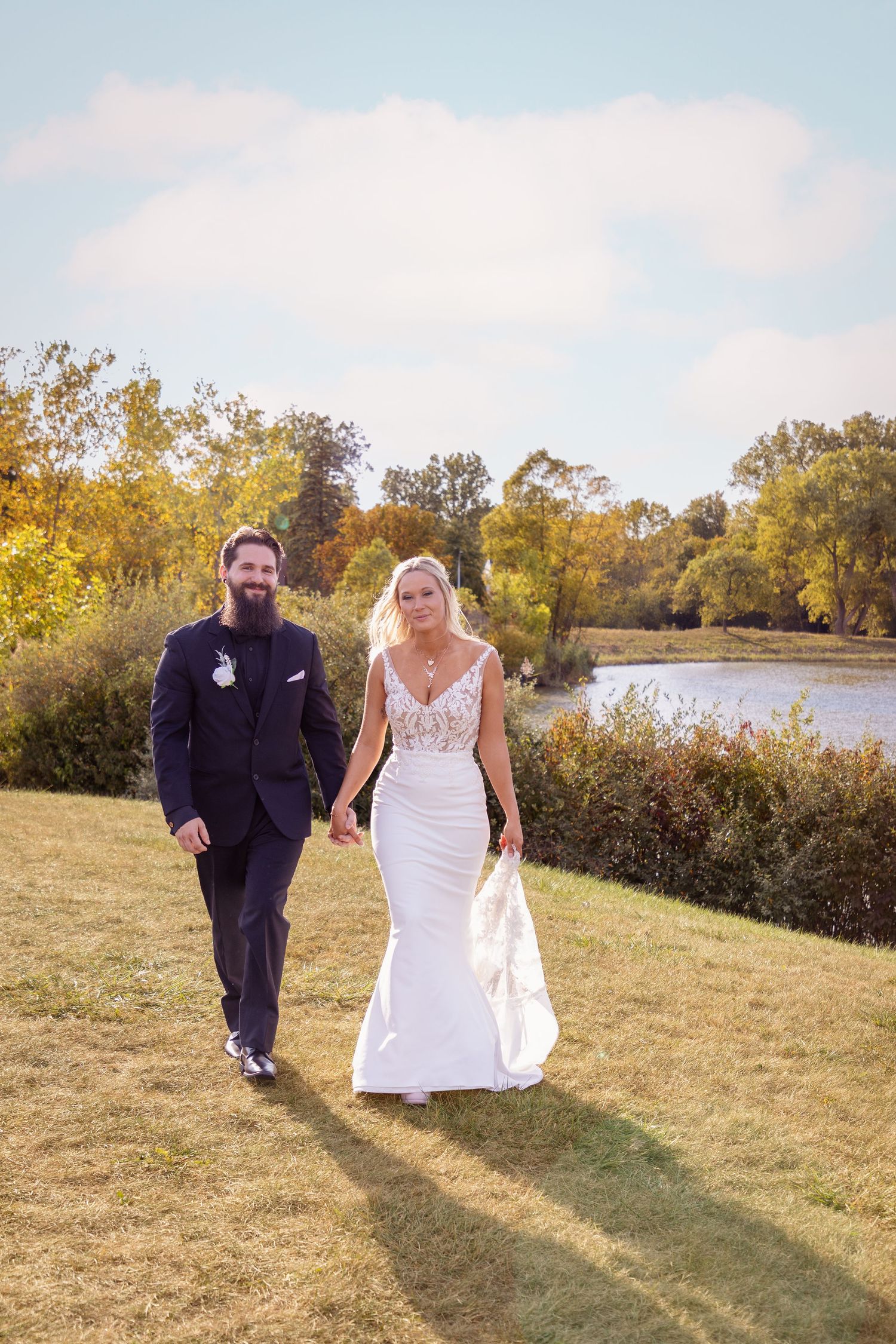 Newlyweds walk hand in hand along a lakeside path on their wedding day.