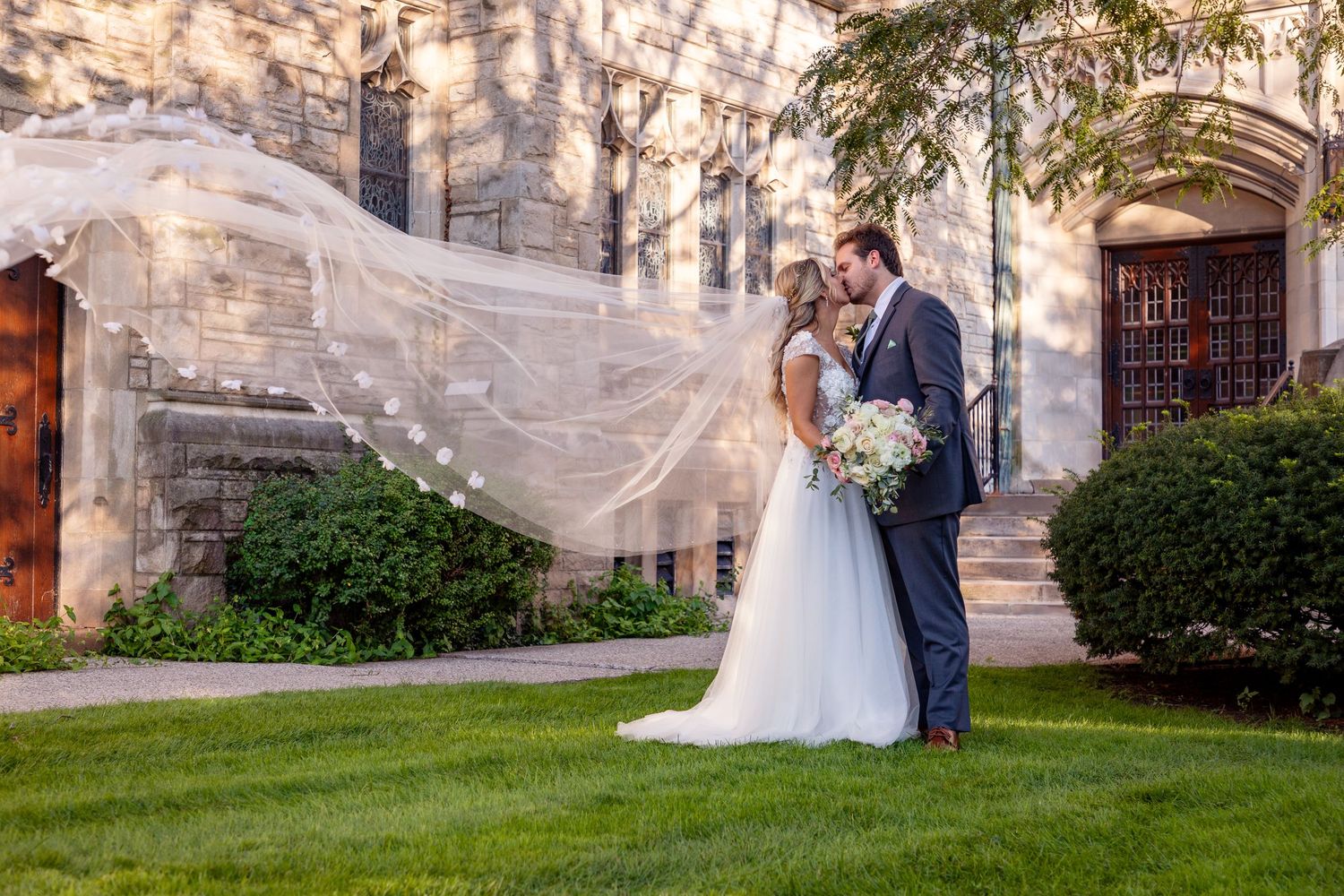 Couple poses romantically with flowing veil against stone church architecture.