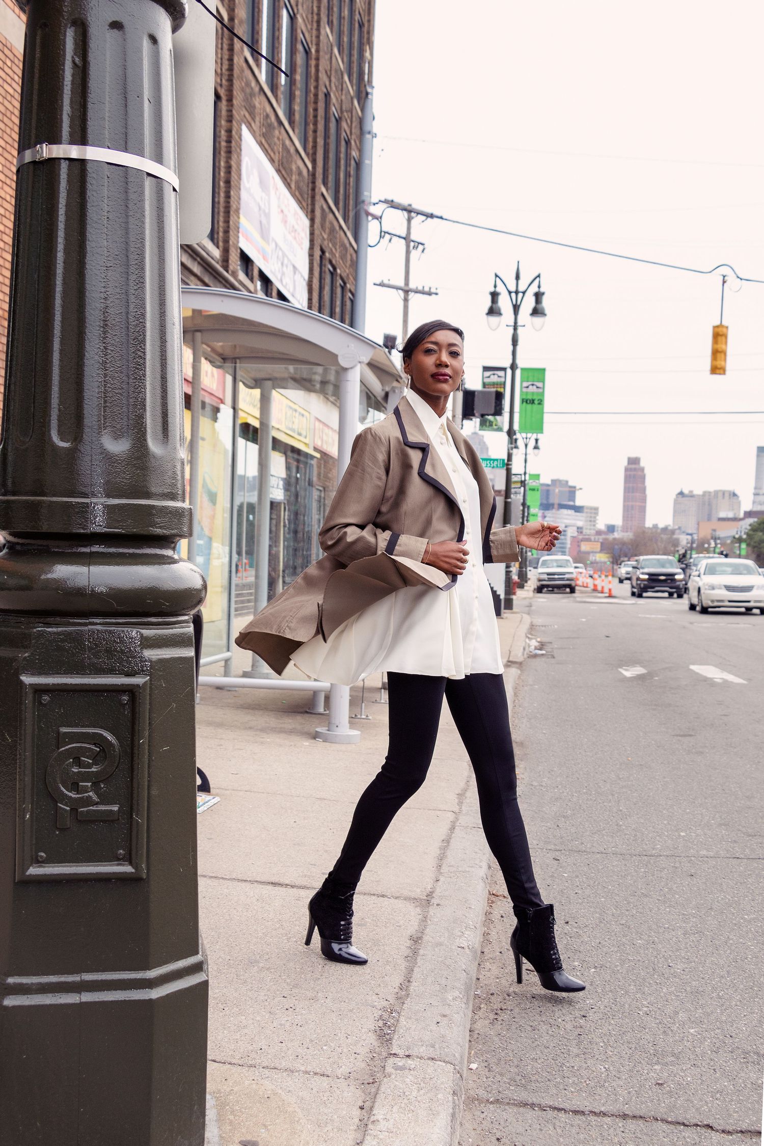 Model in beige blazer and heels posing on urban street with lamp posts.