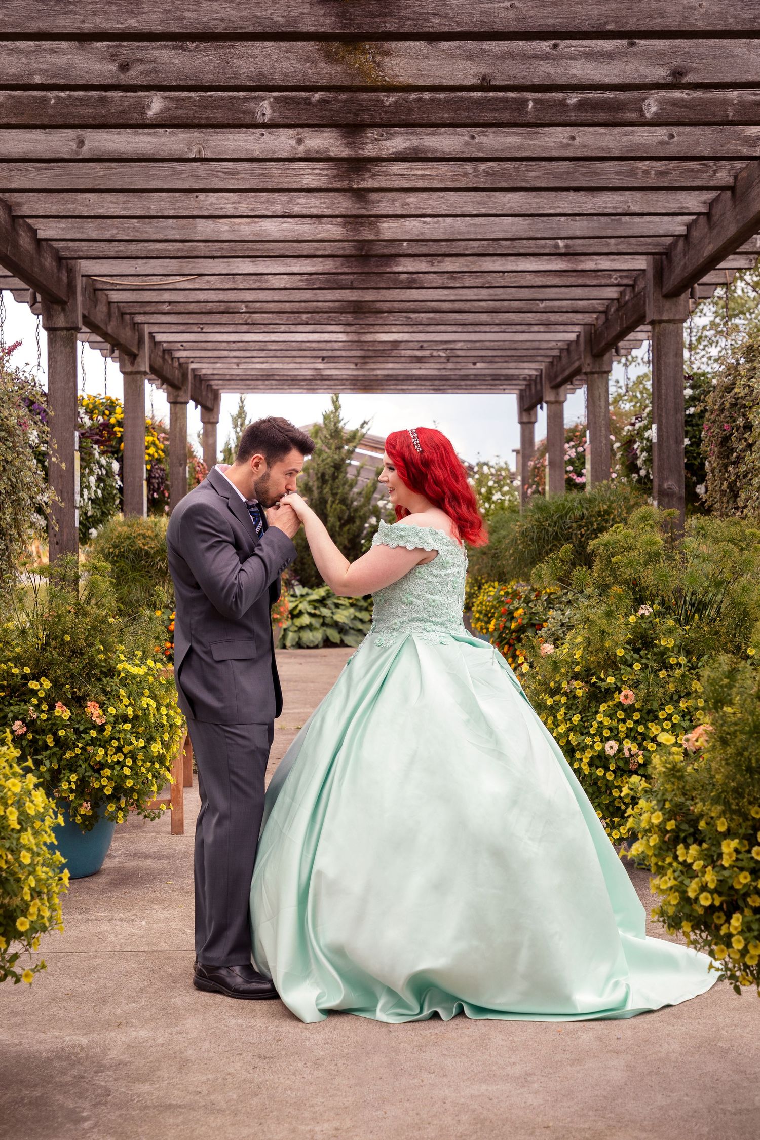 A couple in formal attire poses under a wooden pergola surrounded by yellow flowers.
