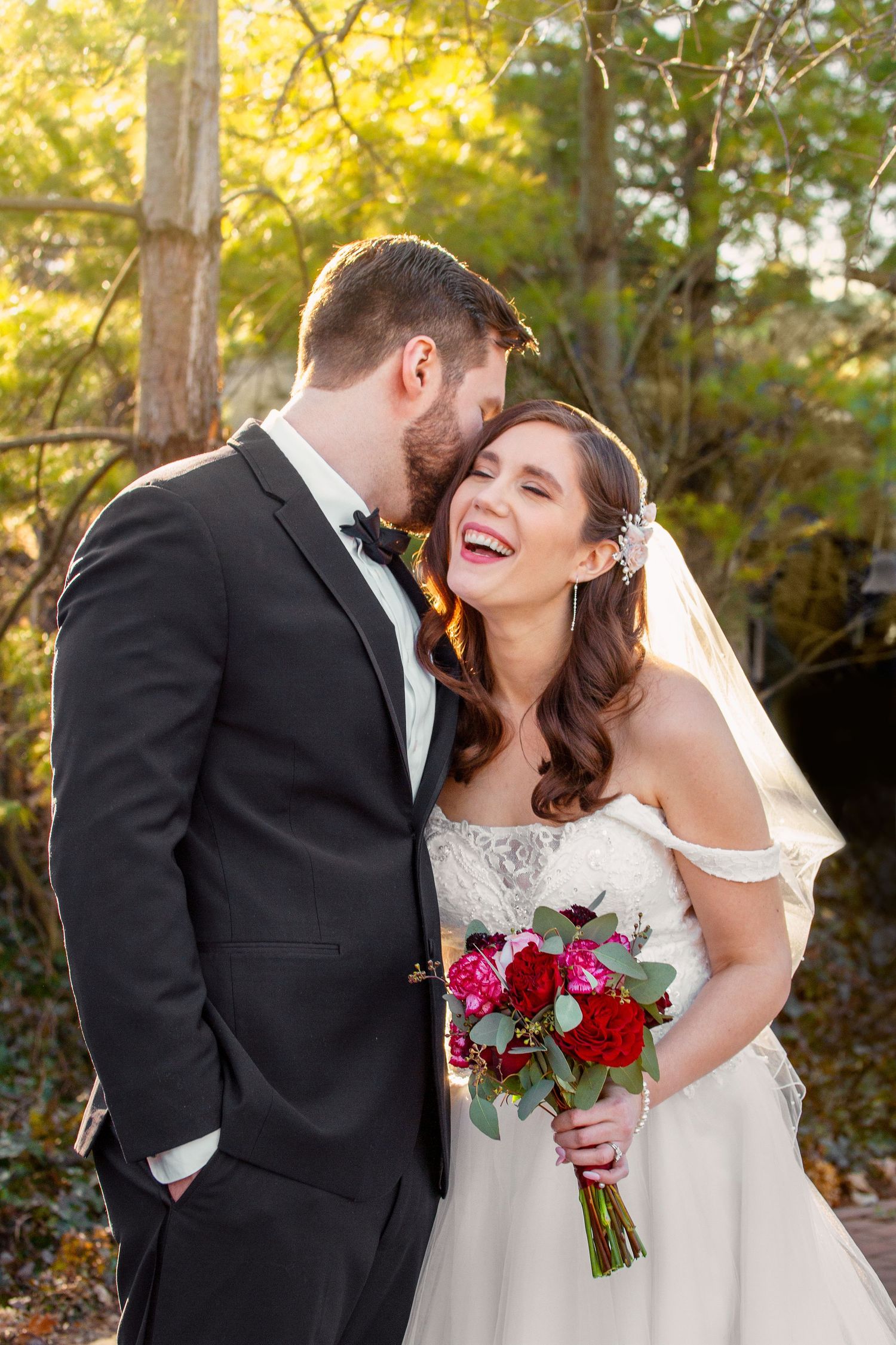 A newlywed couple shares an intimate moment outdoors with the bride holding red roses.
