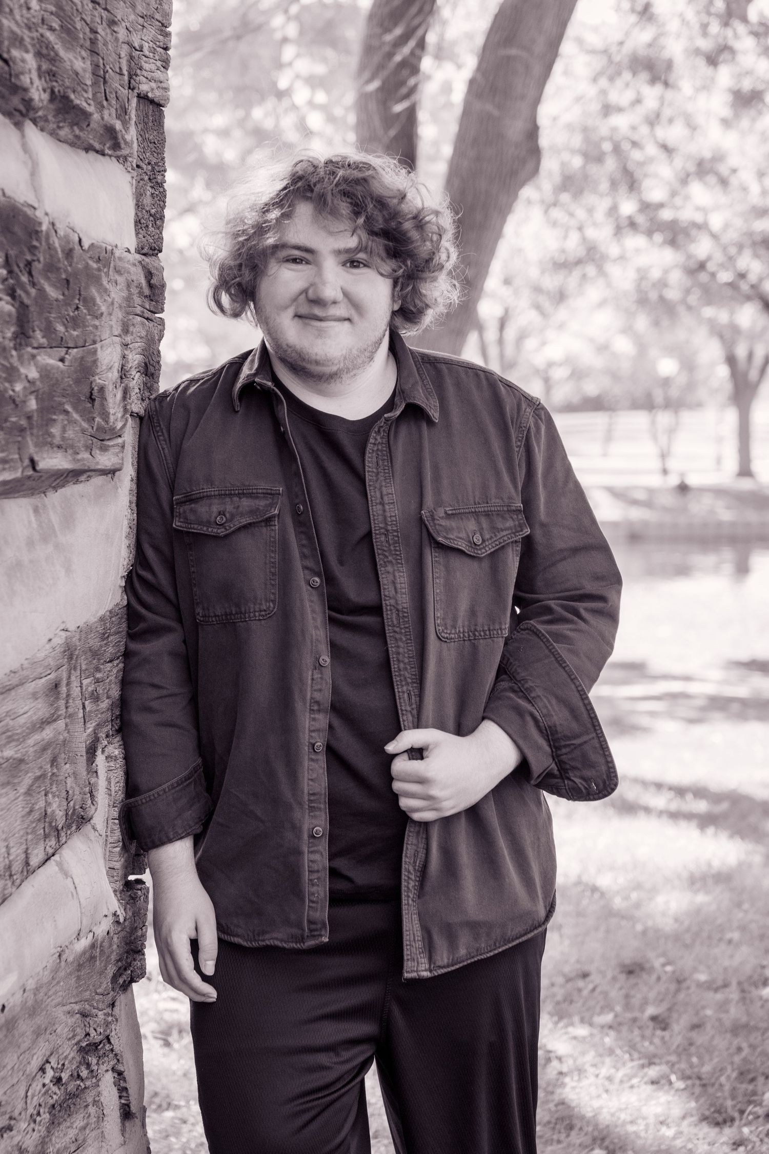 Person in dark denim jacket poses against blurred outdoor background in black and white.