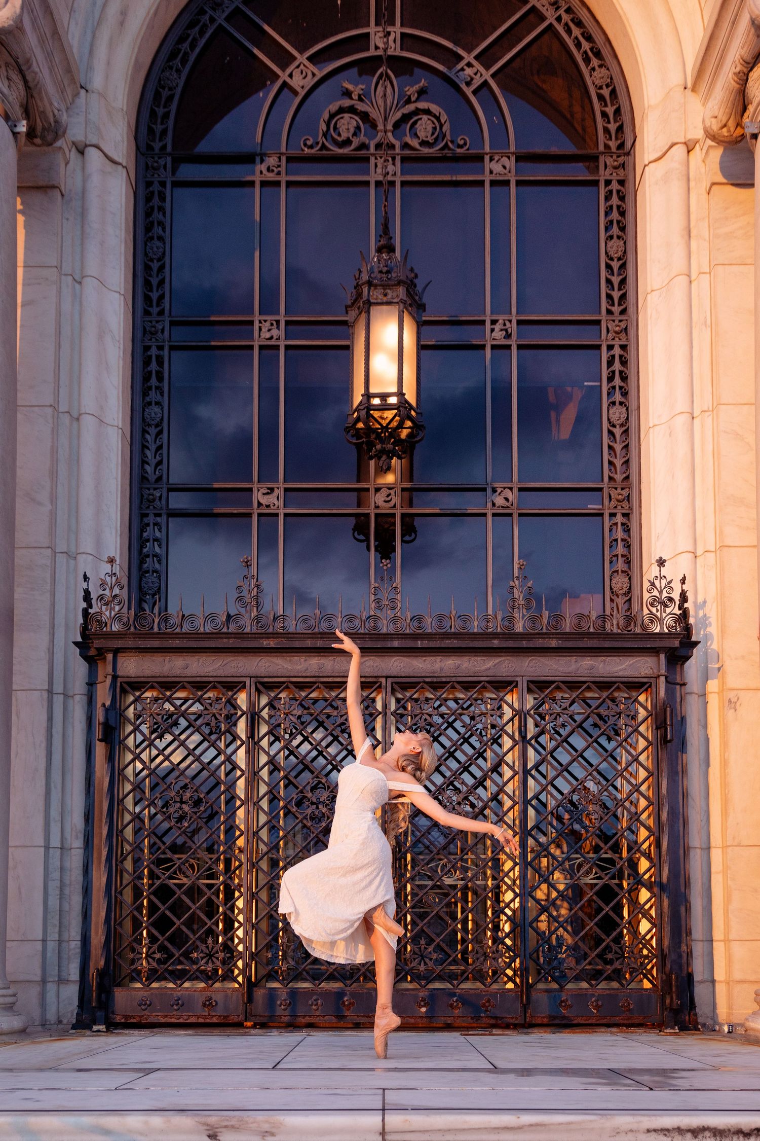 A ballet dancer in a white dress performs an elegant pose against an ornate gothic archway with decorative metalwork.