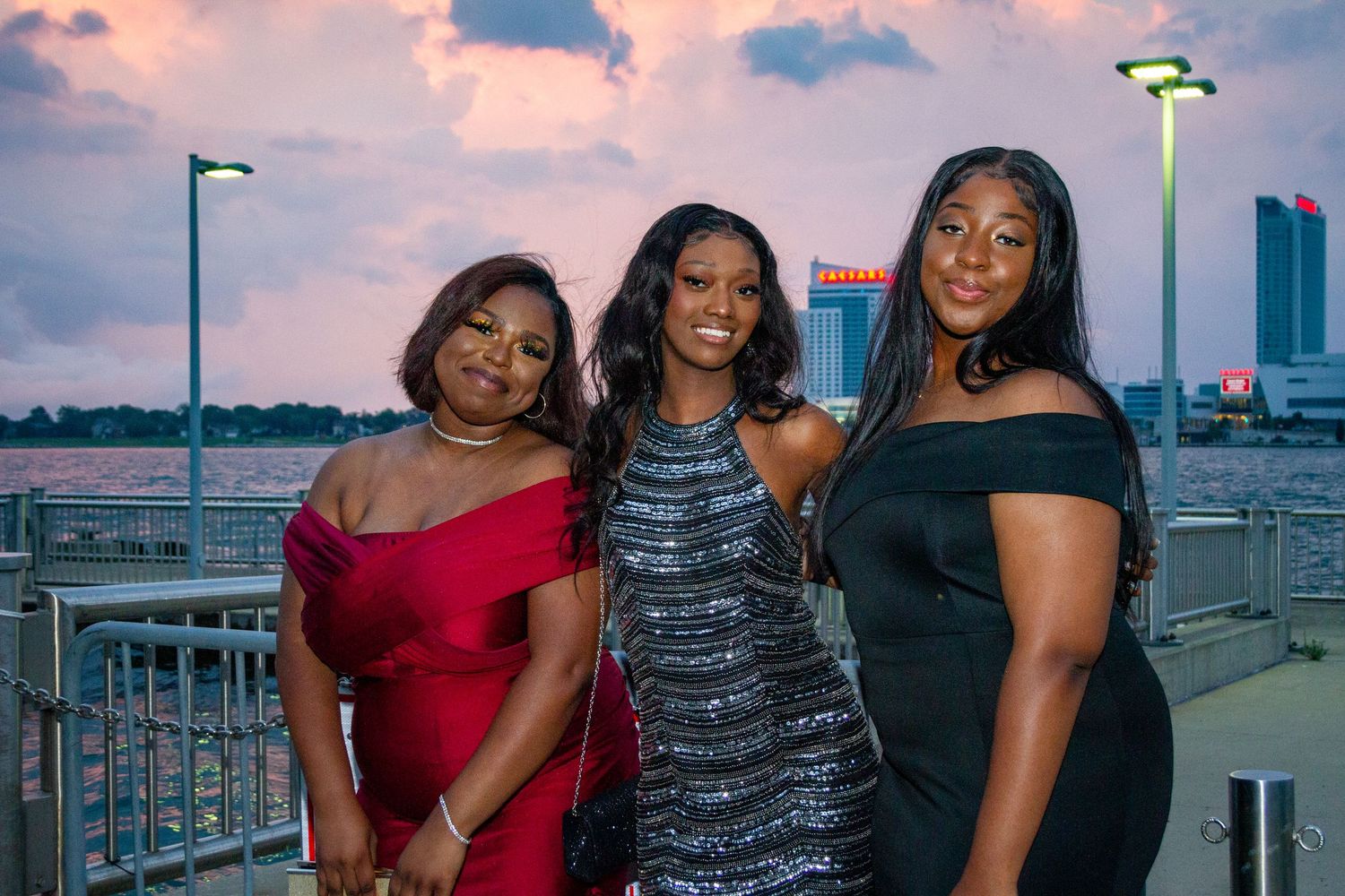Three women in elegant evening wear pose against a sunset cityscape backdrop.