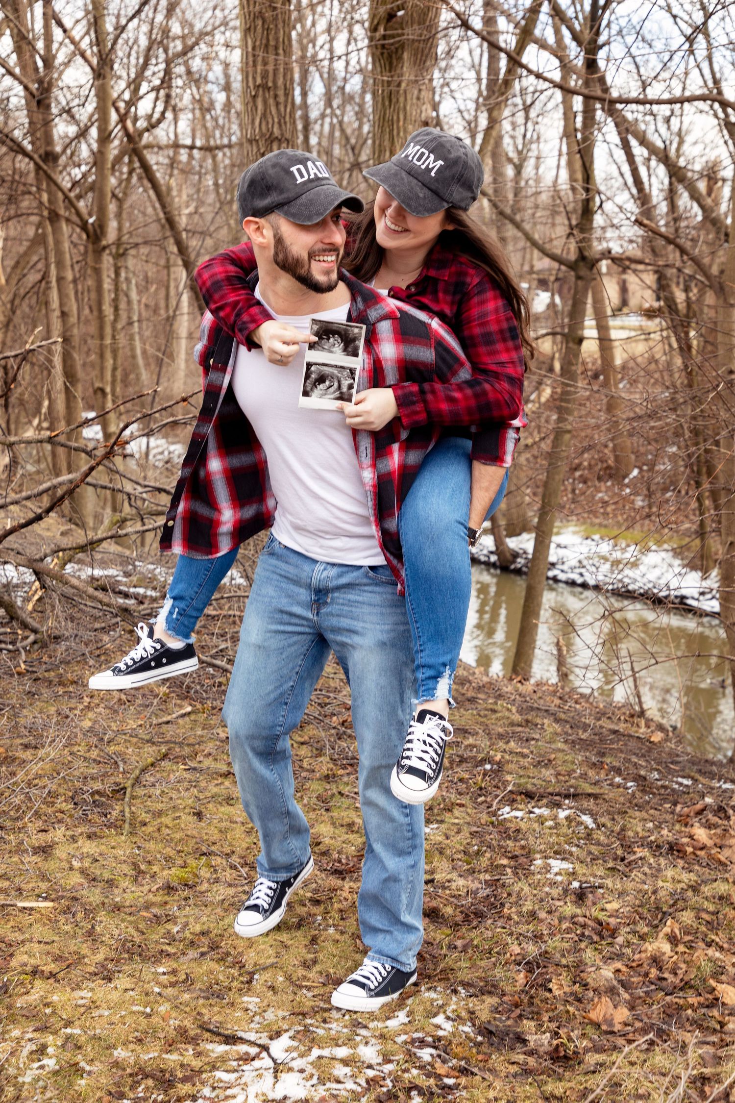 A couple in casual attire walking through a snowy wooded area near a stream.