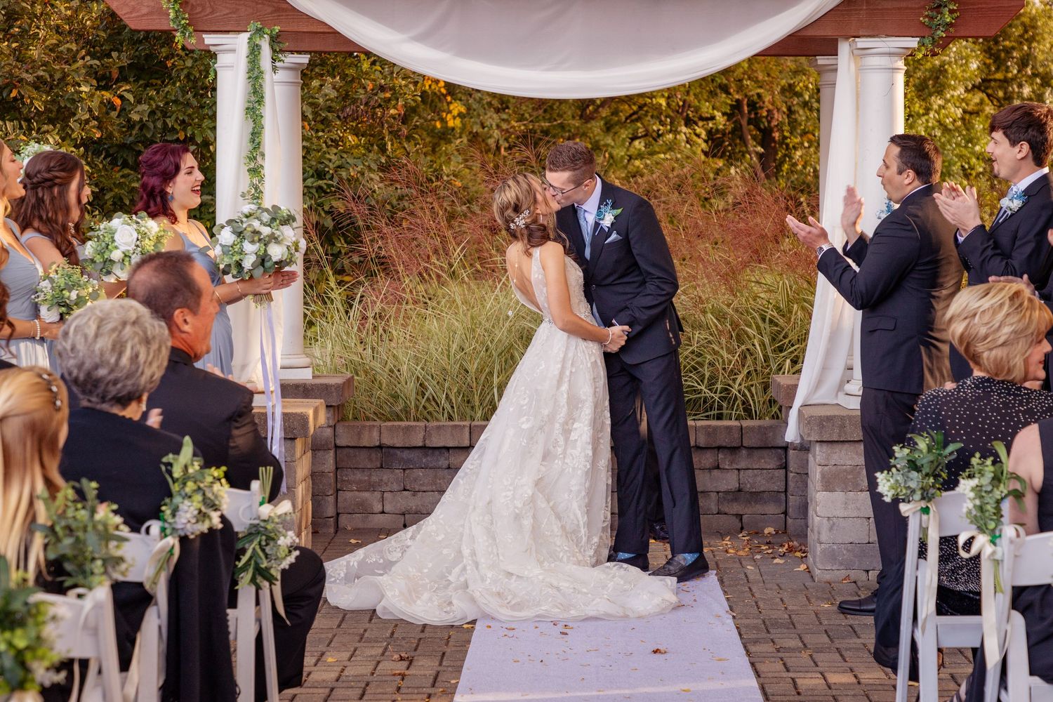 Outdoor wedding ceremony under white draped pergola with guests seated on white chairs.