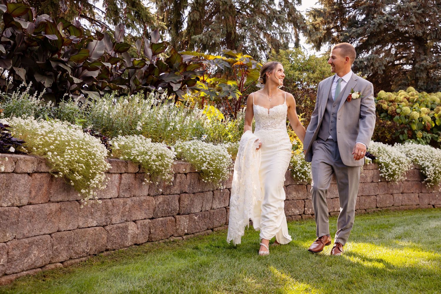 Wedding couple walks along stone wall garden path in afternoon sunlight.