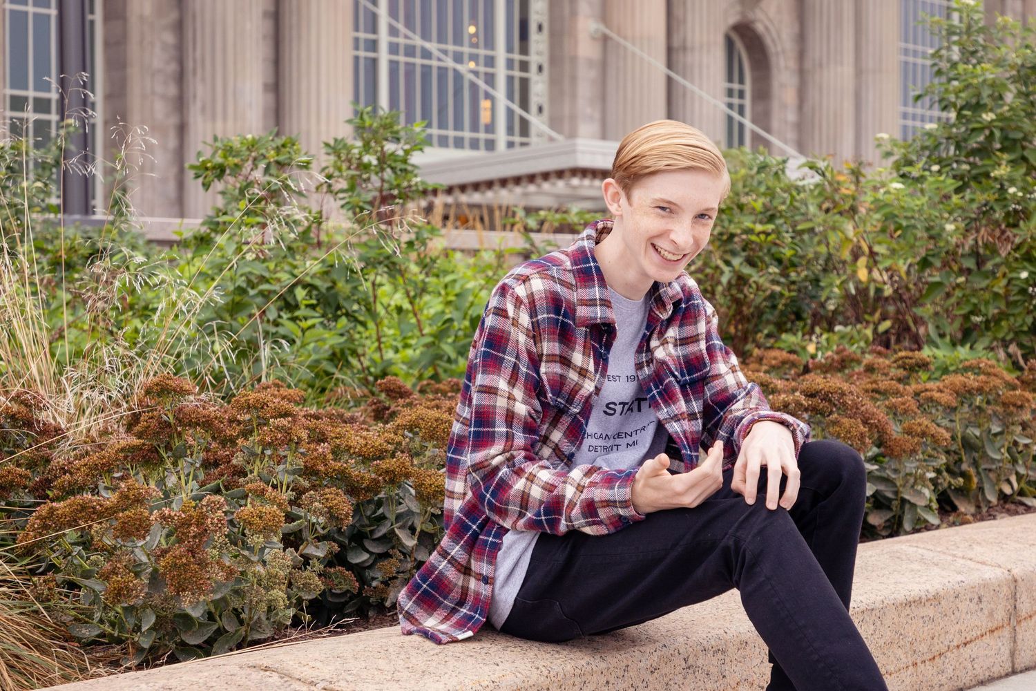 Person in plaid shirt sits casually on stone wall near architectural columns.