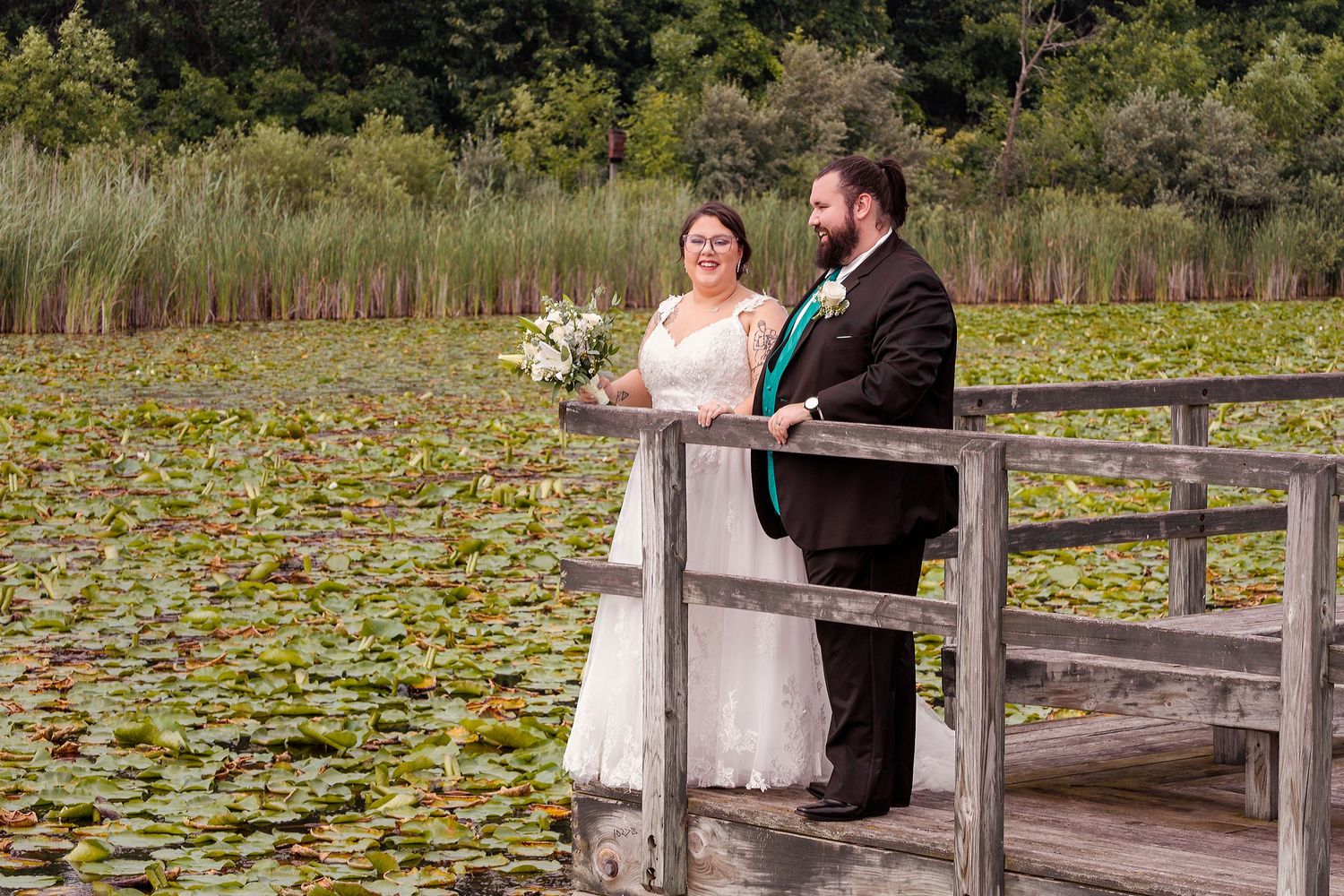 A couple in wedding attire stands on a deck overlooking a lily pond.