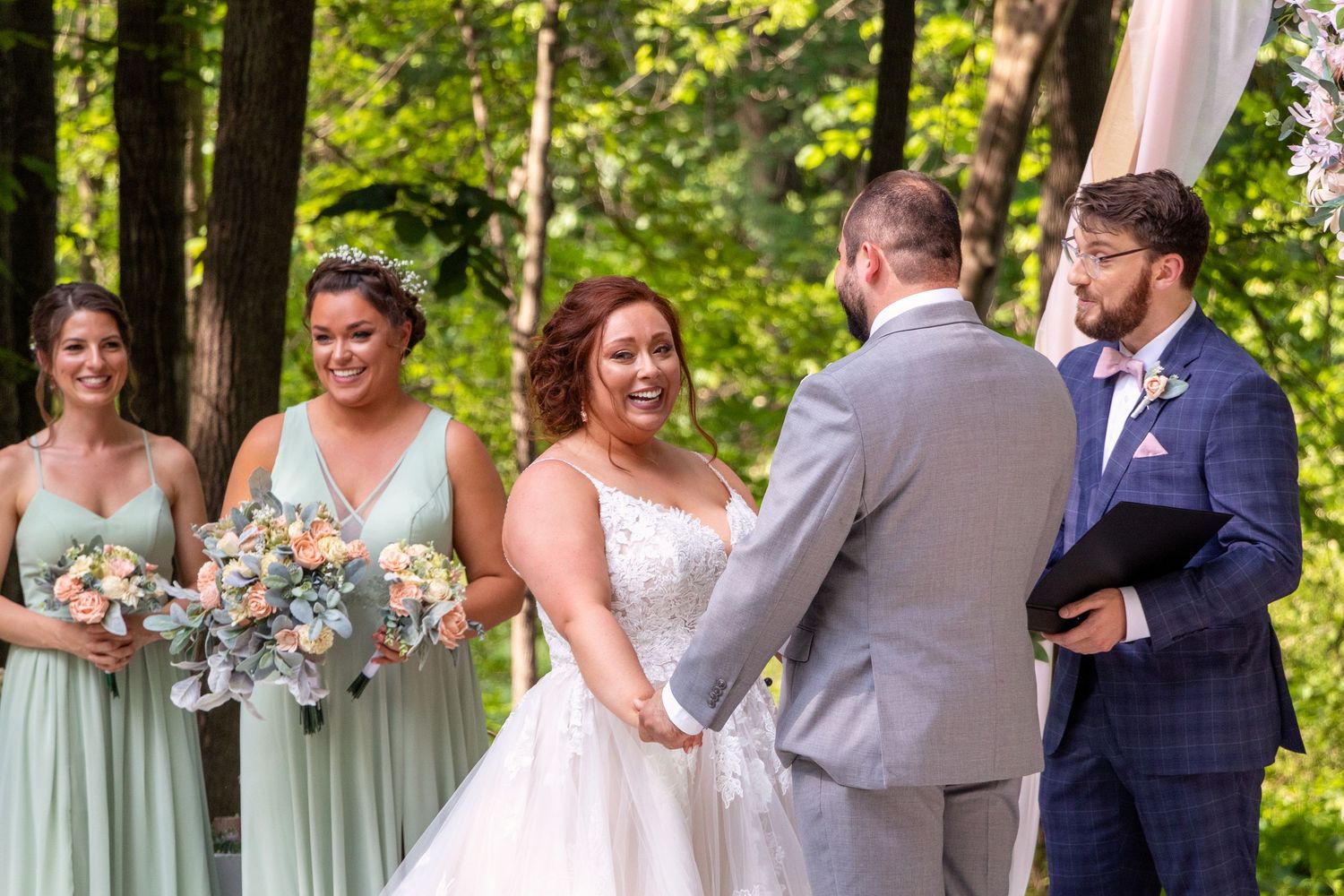 Outdoor wedding ceremony under trees with bridesmaids in mint green dresses watching the exchange of vows.